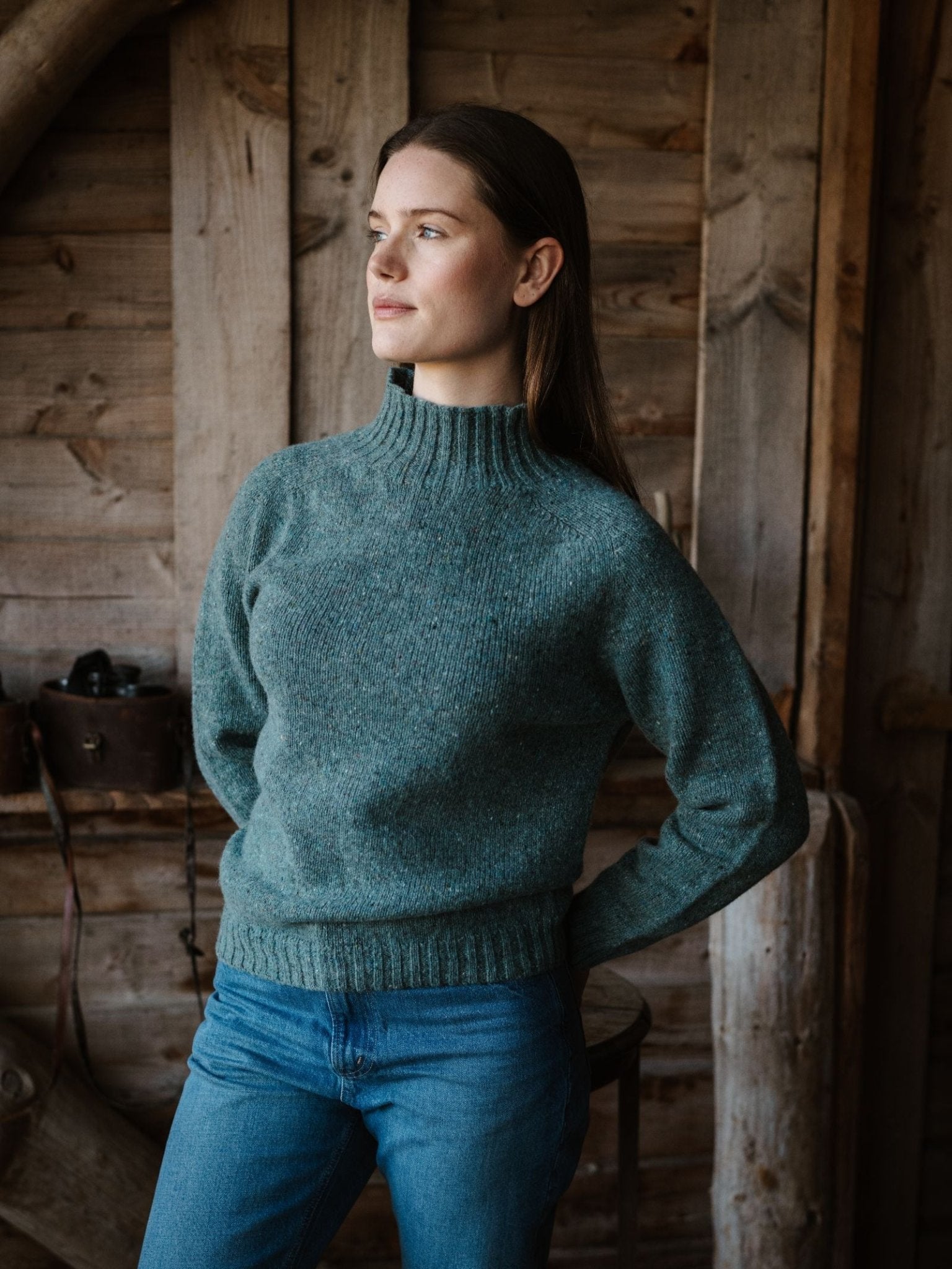 A woman with long brown hair, wearing a teal Campbell’s of Beauly Donegal Lambswool Funnel Neck Jumper and blue jeans, stands indoors against a rustic wooden wall, gazing thoughtfully to the side.