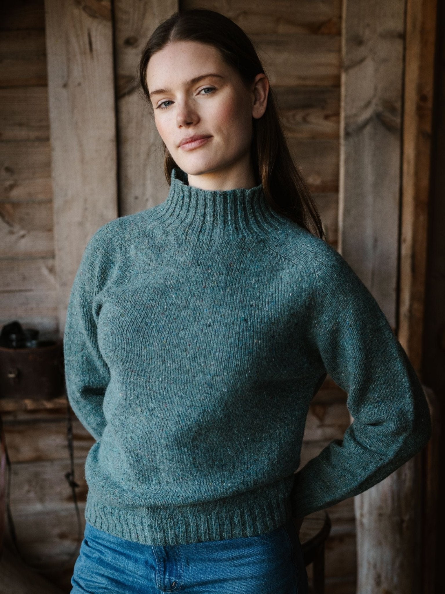 A woman with long brown hair stands indoors against a wooden wall, wearing the Campbell's of Beauly Donegal Lambswool Funnel Neck Jumper in teal and blue jeans, looking calm with her hands behind her back.