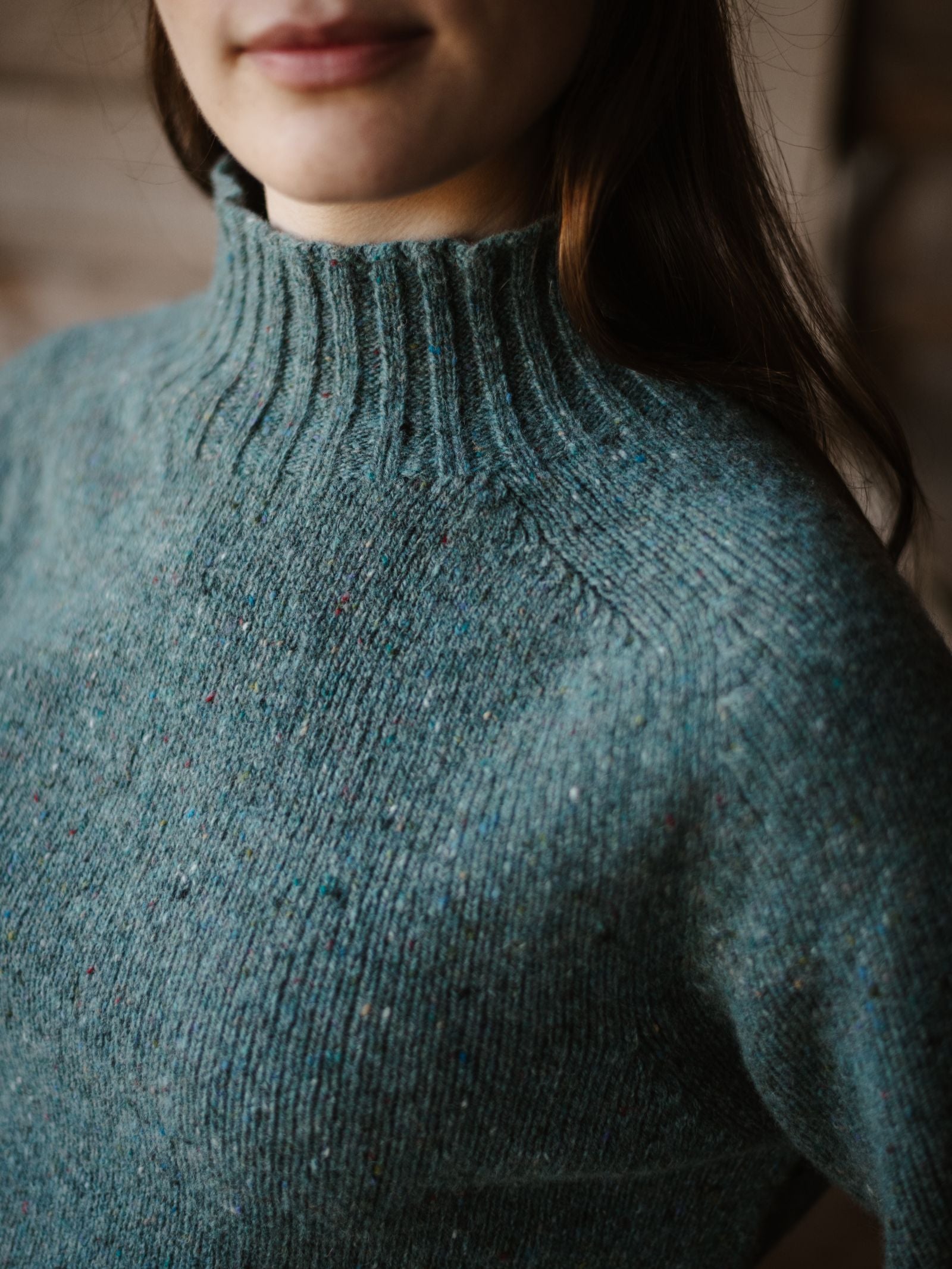 A woman, seen from the shoulders up with a gentle smile, wears a teal Donegal Lambswool Funnel Neck Jumper by Campbell's of Beauly. The background is blurred.