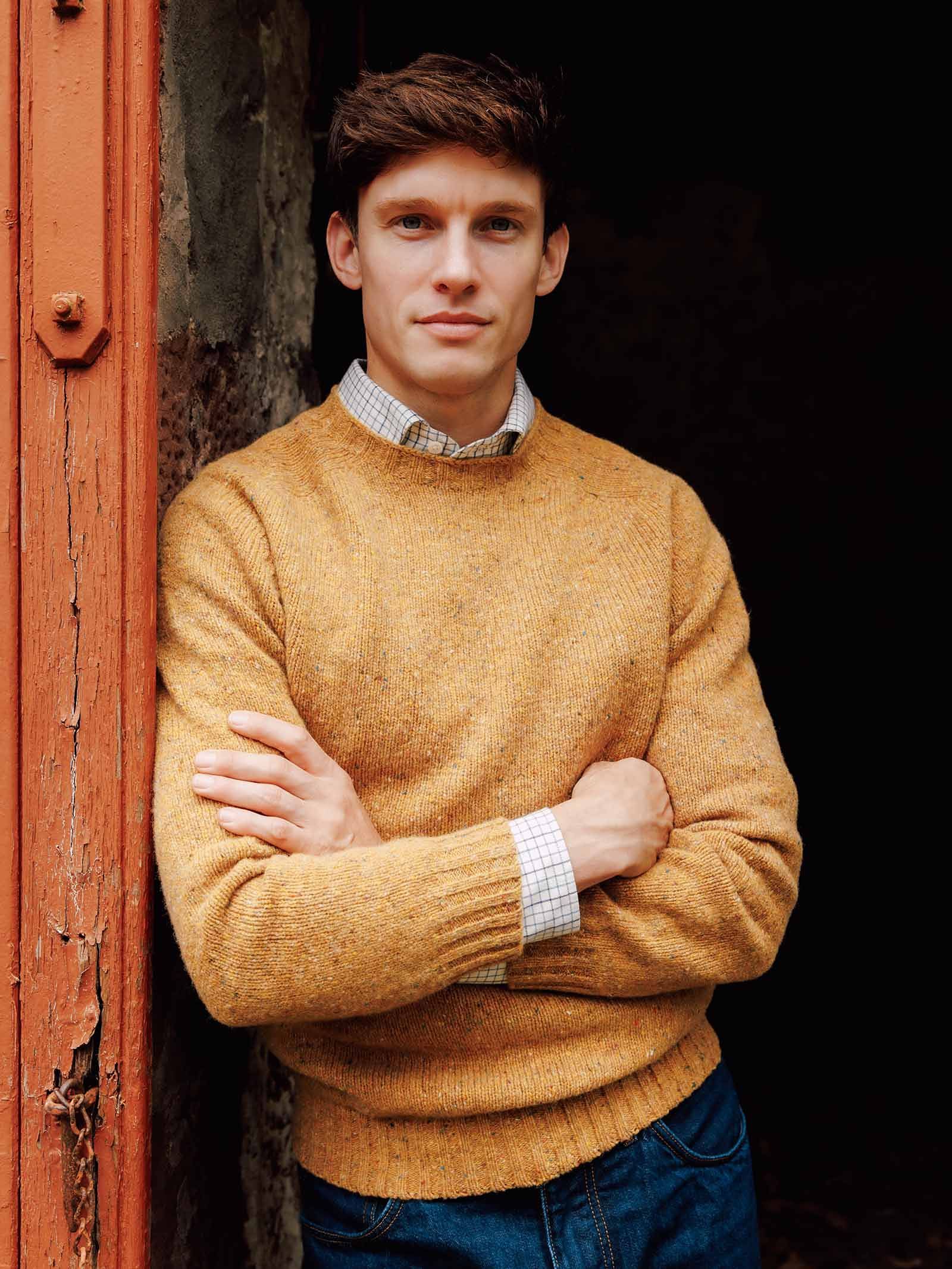 A man stands with his arms crossed, wearing the Campbell’s of Beauly Donegal Lambswool Crew Neck Jumper in mustard yellow over a checked shirt and blue jeans, leaning against a weathered red wooden doorframe.