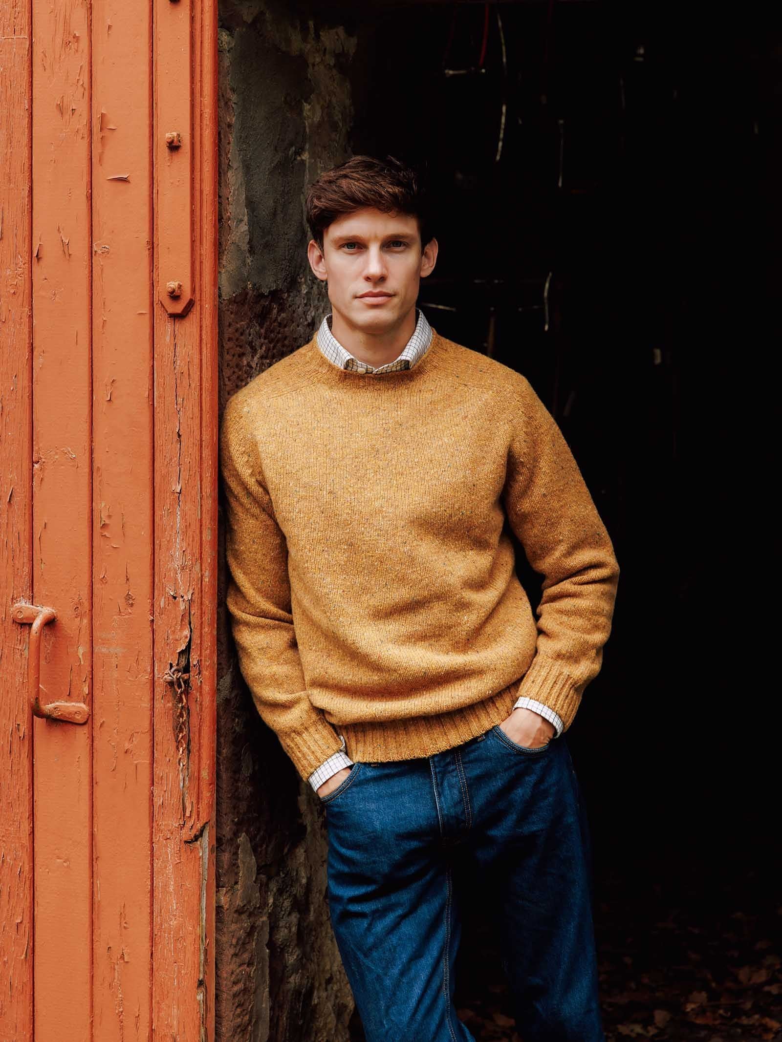 A man in a mustard-yellow Donegal Lambswool Crew Neck Jumper from Campbell’s of Beauly, crafted in Scotland from soft lambswool, stands leaning against an orange wooden doorframe, wearing blue jeans and looking at the camera with his hands in his pockets. The background is dark and rustic.