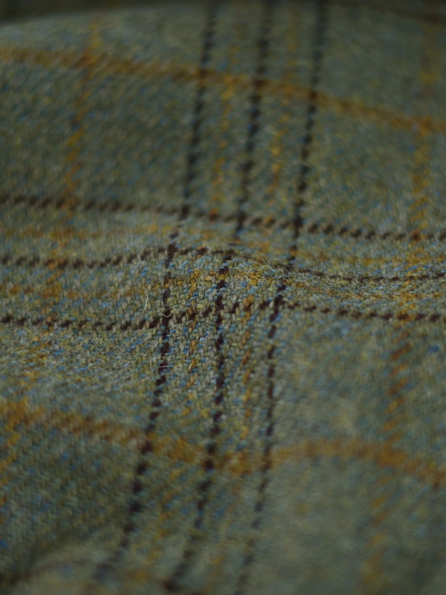Close-up of green and brown plaid fabric, showing a textured wool surface with intersecting lines forming a checkered pattern. The image highlights the detailed weave and fibers of the material.