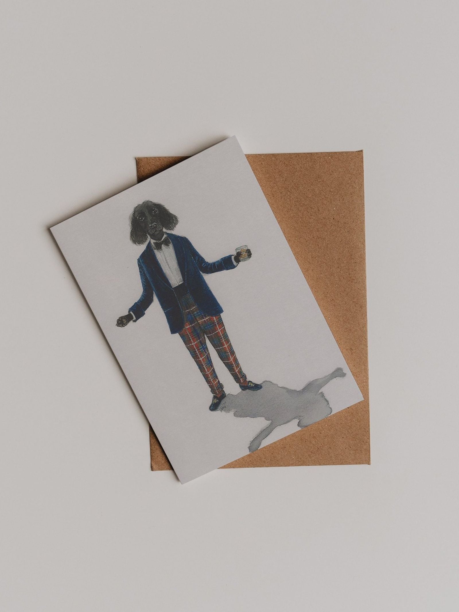 The Evening Wear Greeting Card by Campbell’s of Beauly showcases a dog dressed in a suit and plaid pants standing on a shadow, set atop a brown envelope against a white background—a playful and elegant thank you card designed in Sophie Murray’s charming style, perfect for any occasion.