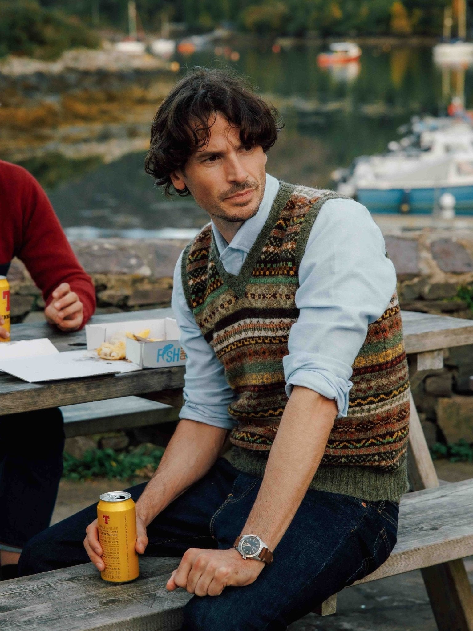 A man in a Campbell's of Beauly Fairisle Slipover over a light blue shirt sits on a wooden bench outdoors, holding a can of beer, with water, boats, and another person beside him against a stone wall in the background.