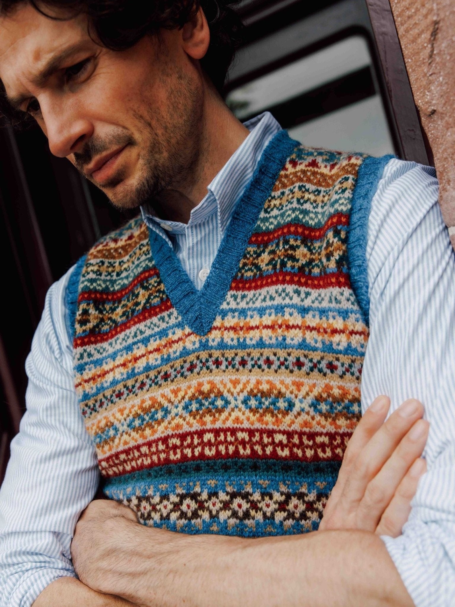 A man in a blue and white striped shirt wears the Campbell's of Beauly Fairisle Slipover. With arms crossed by a window and stone wall, he thoughtfully showcases this classic sleeveless knit.