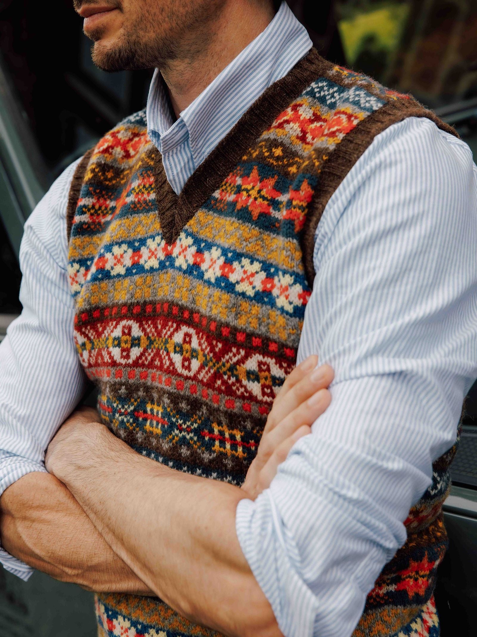 A person with arms crossed wears a blue striped shirt and the Campbell’s of Beauly Fairisle Slipover—a sleeveless knit layer crafted from soft Shetland wool, featuring colorful geometric patterns.