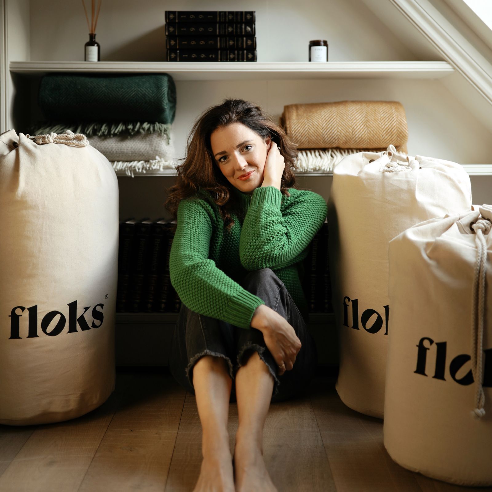 A woman in a green sweater sits barefoot on the floor between two large canvas bags labeled floks, with folded blankets and books on shelves behind her.
