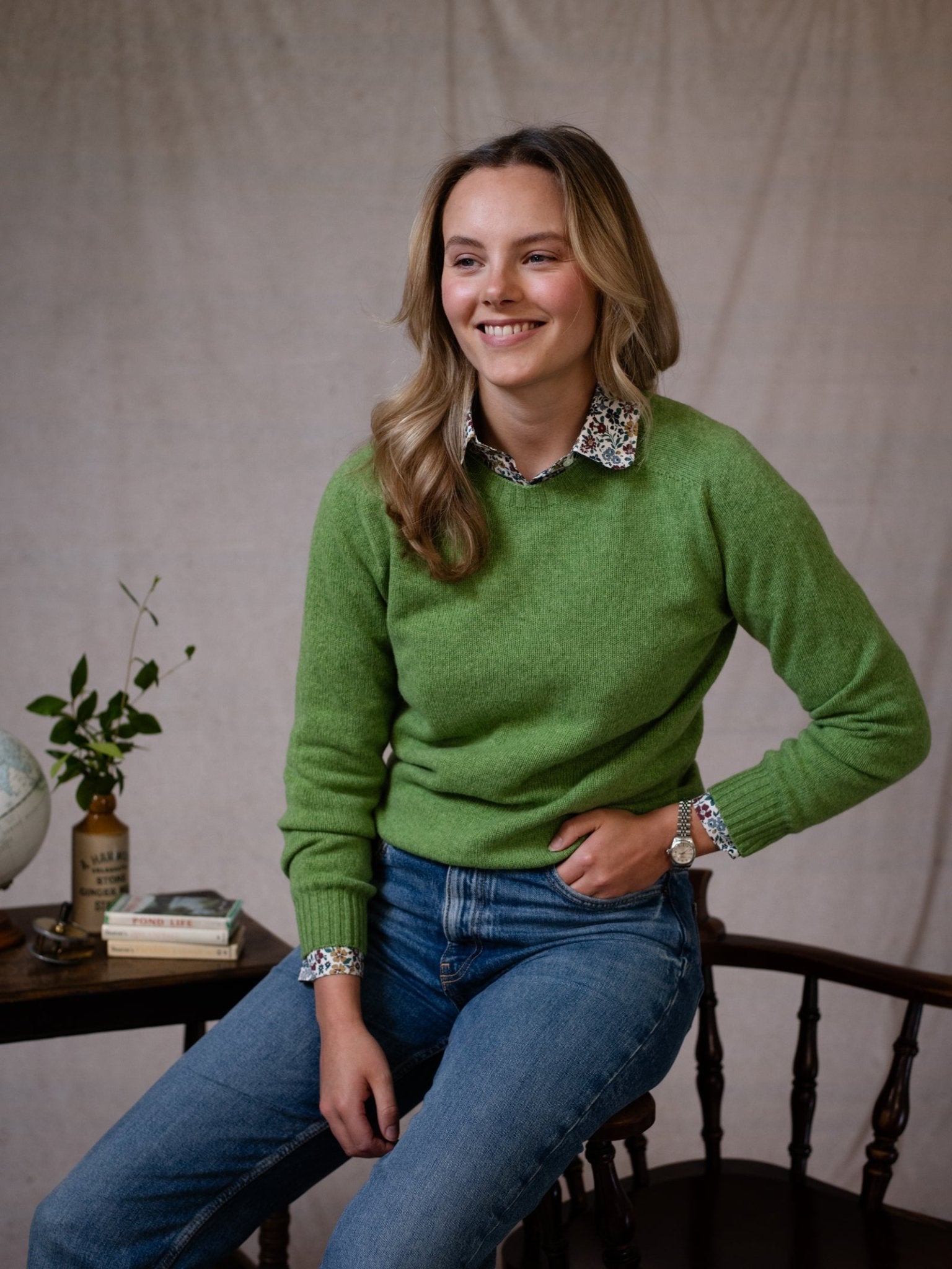 A blonde woman in blue jeans and a green Geelong Lambswool Crew Neck Jumper by Campbell’s of Beauly sits smiling on a wooden chair. Beside her, a table holds a potted plant, globe, and books. Neutral fabric backdrop. Made in Scotland.