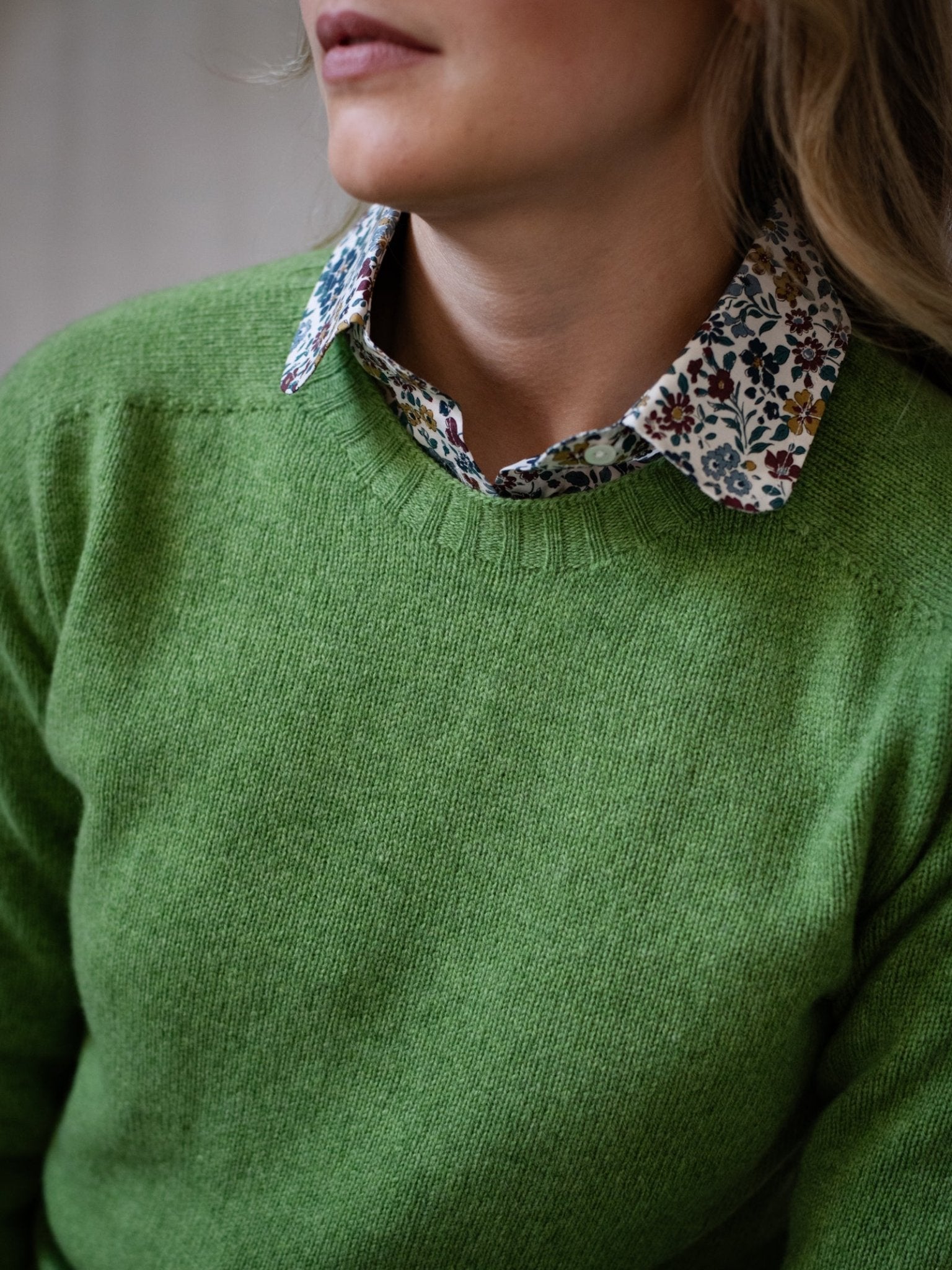 A person wears a green Campbell's of Beauly Geelong Lambswool Crew Neck Jumper over a floral-collared shirt. The cropped photo focuses on the torso and face, highlighting the Scottish-made clothing details.