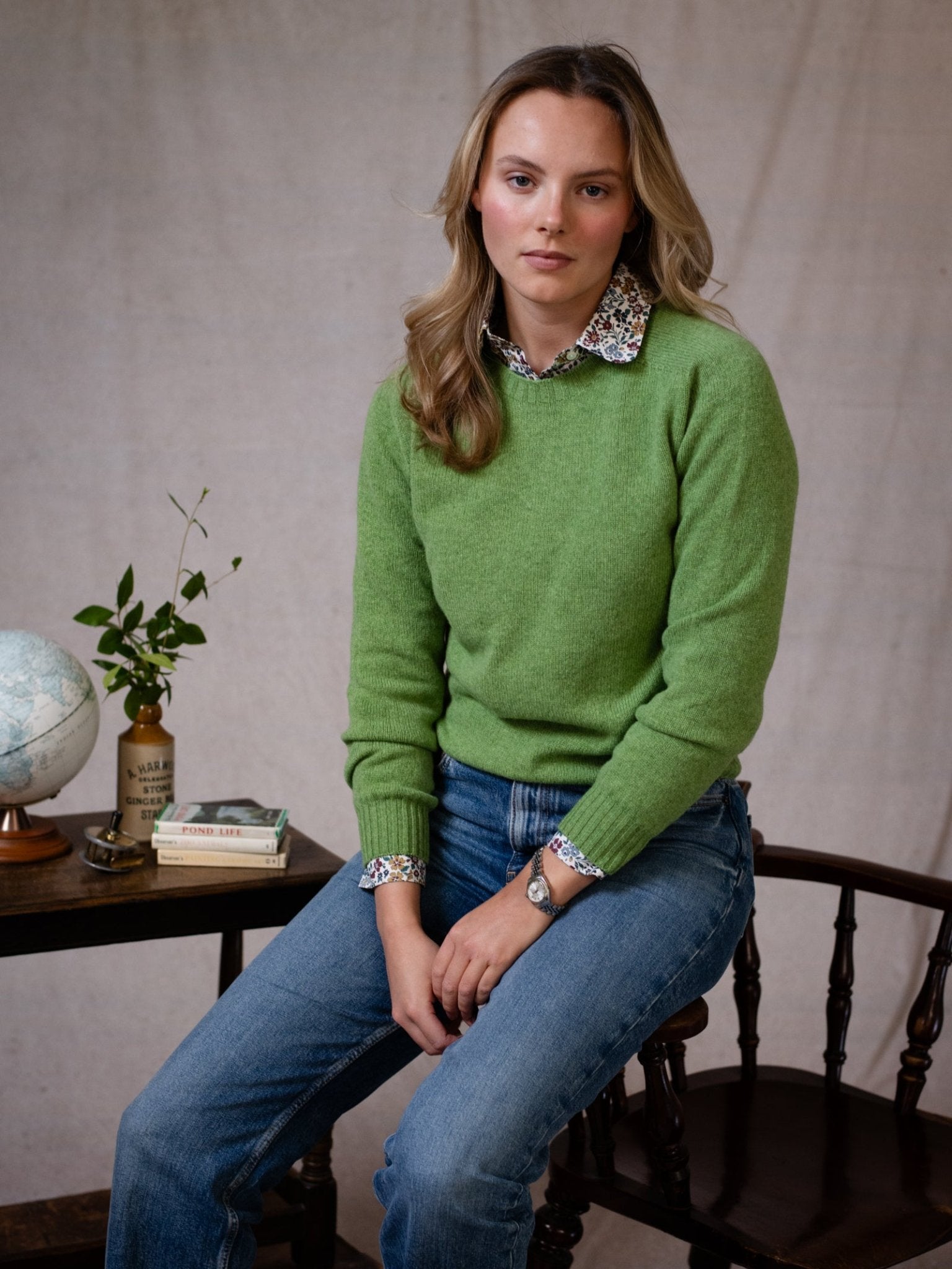 A woman with long blonde hair sits on a wooden chair, wearing Campbell's of Beauly Geelong Lambswool Crew Neck Jumper in green over a floral collared shirt and blue jeans. A table nearby holds a globe, books, a plant, and vase against a light fabric background.