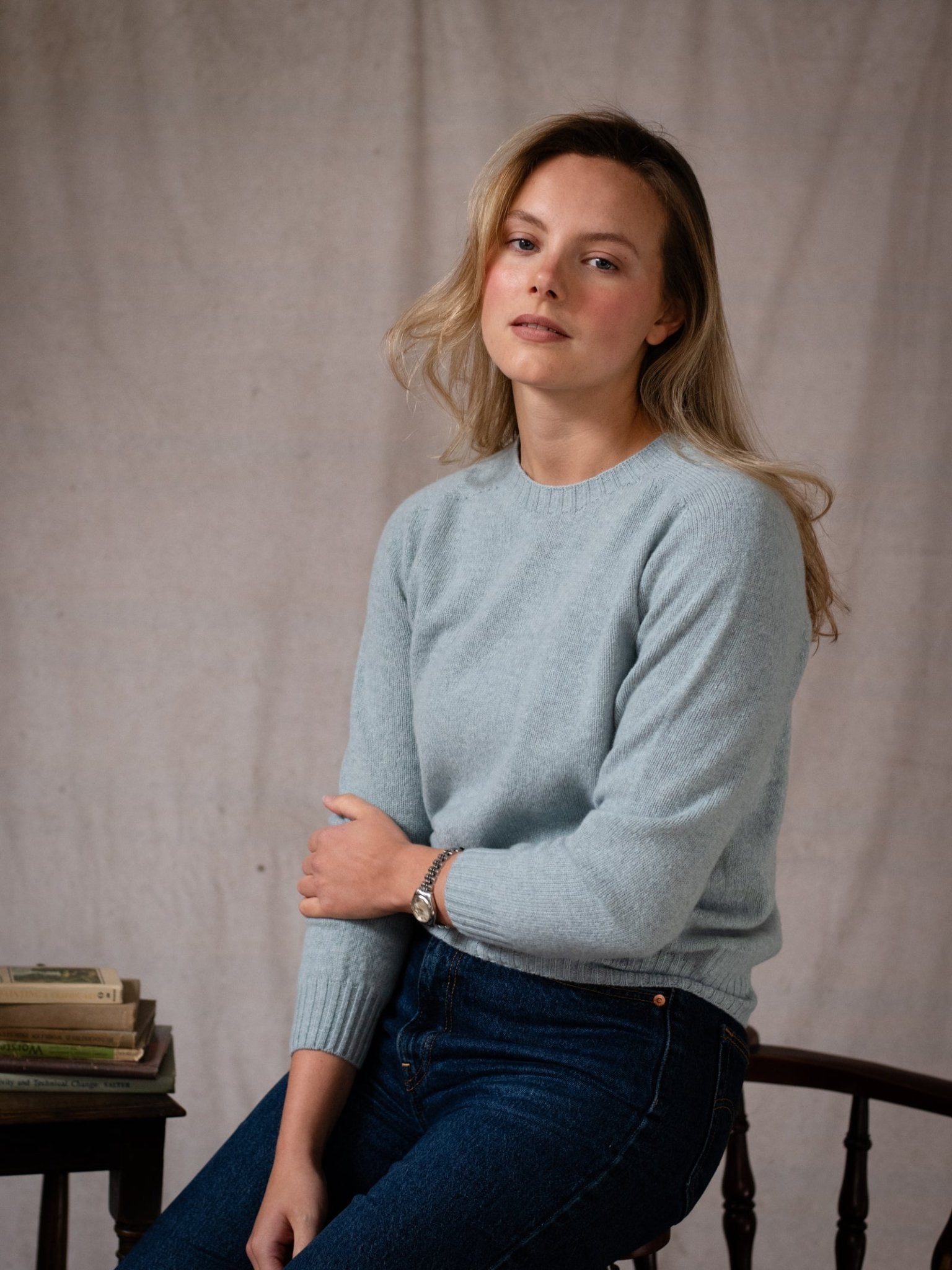 A person with long blonde hair, wearing the Campbell's of Beauly Geelong Lambswool Crew Neck Jumper and jeans, sits on a chair beside a small stack of books against a neutral fabric backdrop.