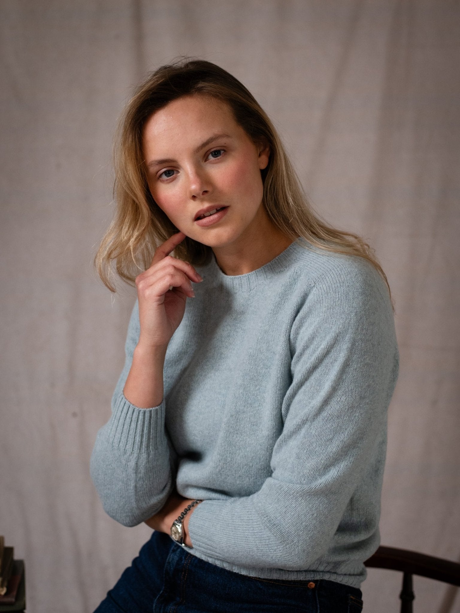 A woman with blonde hair wears the Campbell's of Beauly Geelong Lambswool Crew Neck Jumper in light blue, seated on a chair, resting her elbow on her knee and touching her chin while looking thoughtfully at the camera against a neutral background.