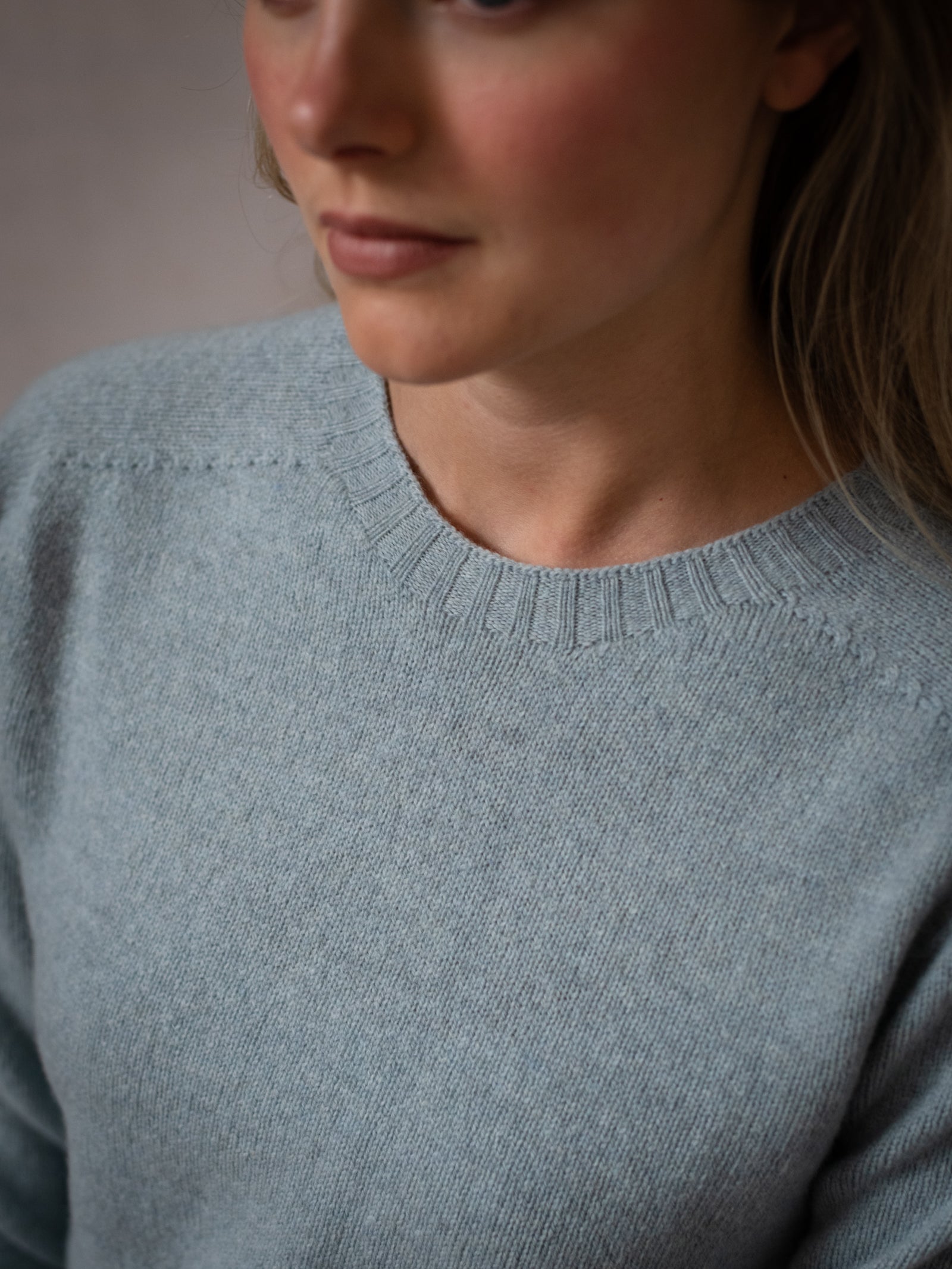 A close-up of a woman in a Campbell's of Beauly Geelong Lambswool Crew Neck Jumper in light greyish-blue, her face partially visible as she gazes to the side against a softly blurred background.