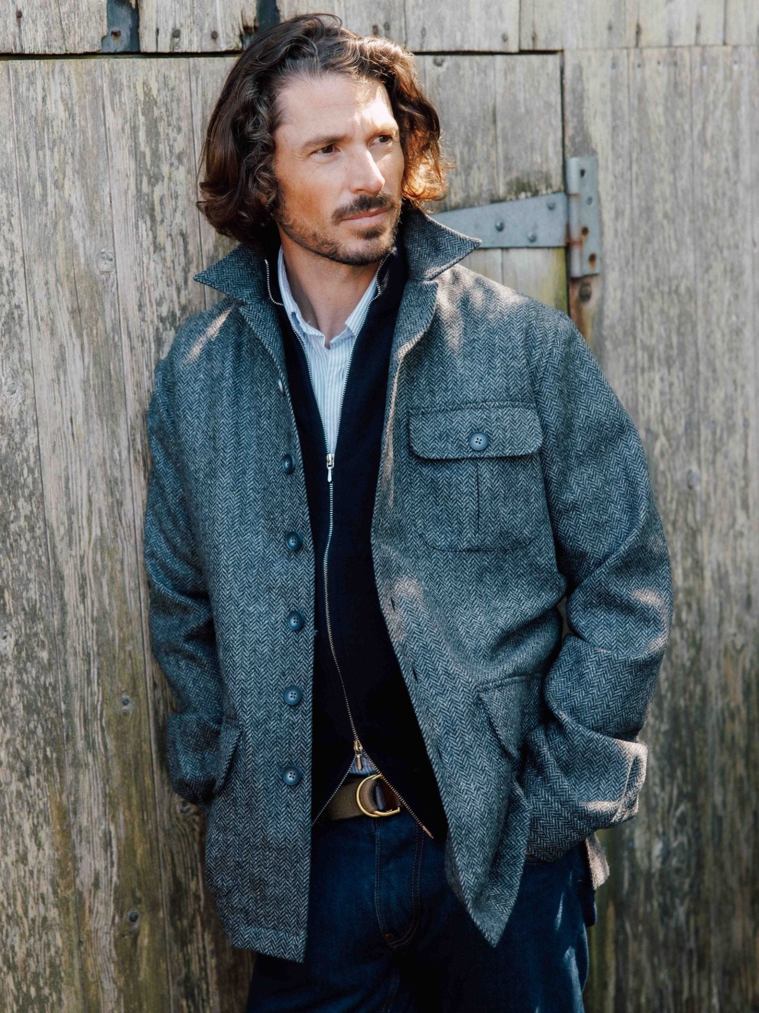 A man with wavy brown hair and a beard stands against a weathered wooden wall wearing the Campbell's of Beauly Casual Tweed Shacket over a dark sweater and jeans, hands in his pockets, embodying relaxed tailoring as he looks to the side.