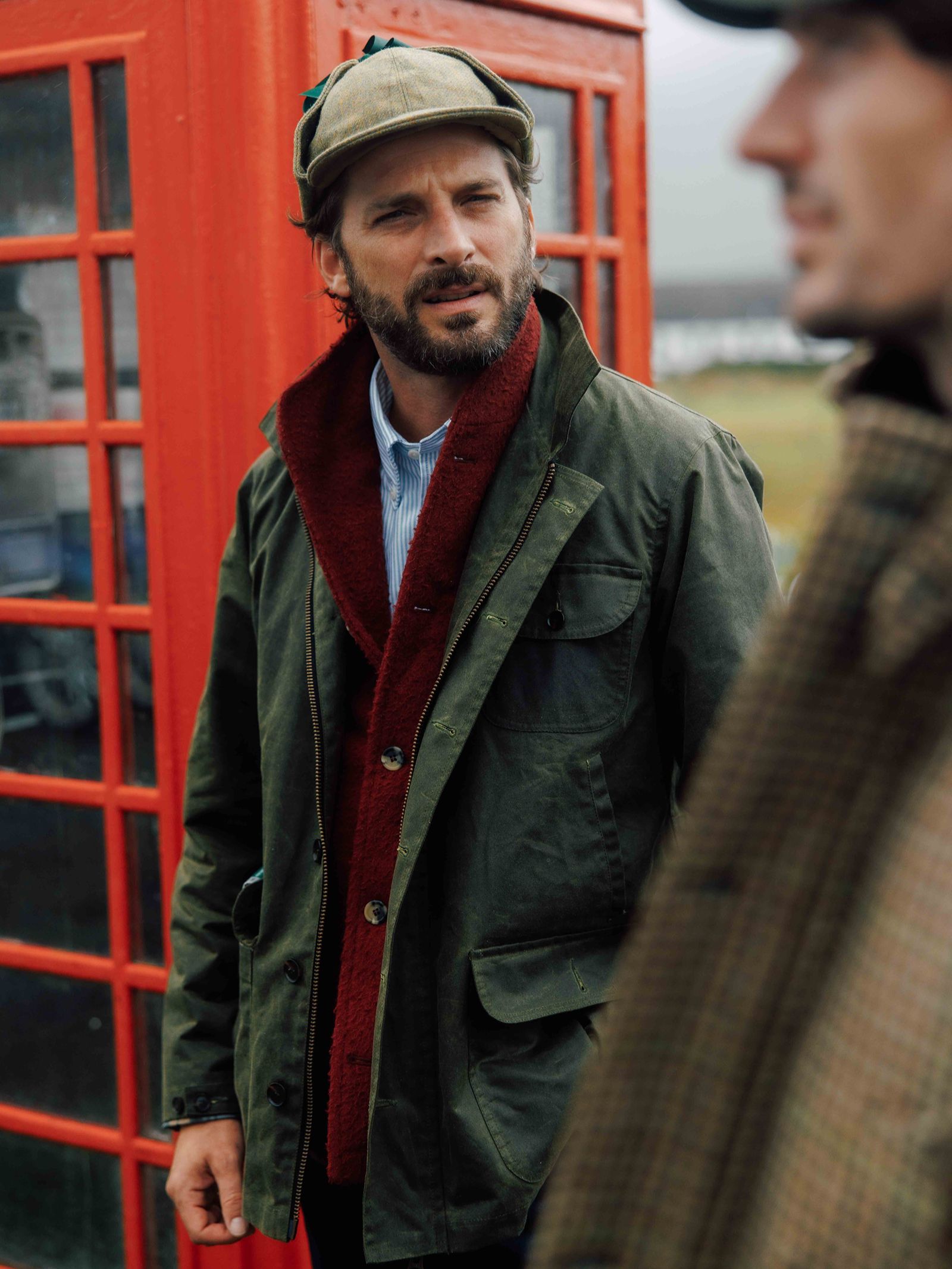 A man wearing the field-ready Garve Jacket in olive green waxed cotton by Campbell’s of Beauly, along with a matching cap, stands near a red phone booth and looks at another person in a plaid coat outdoors on a cloudy day.