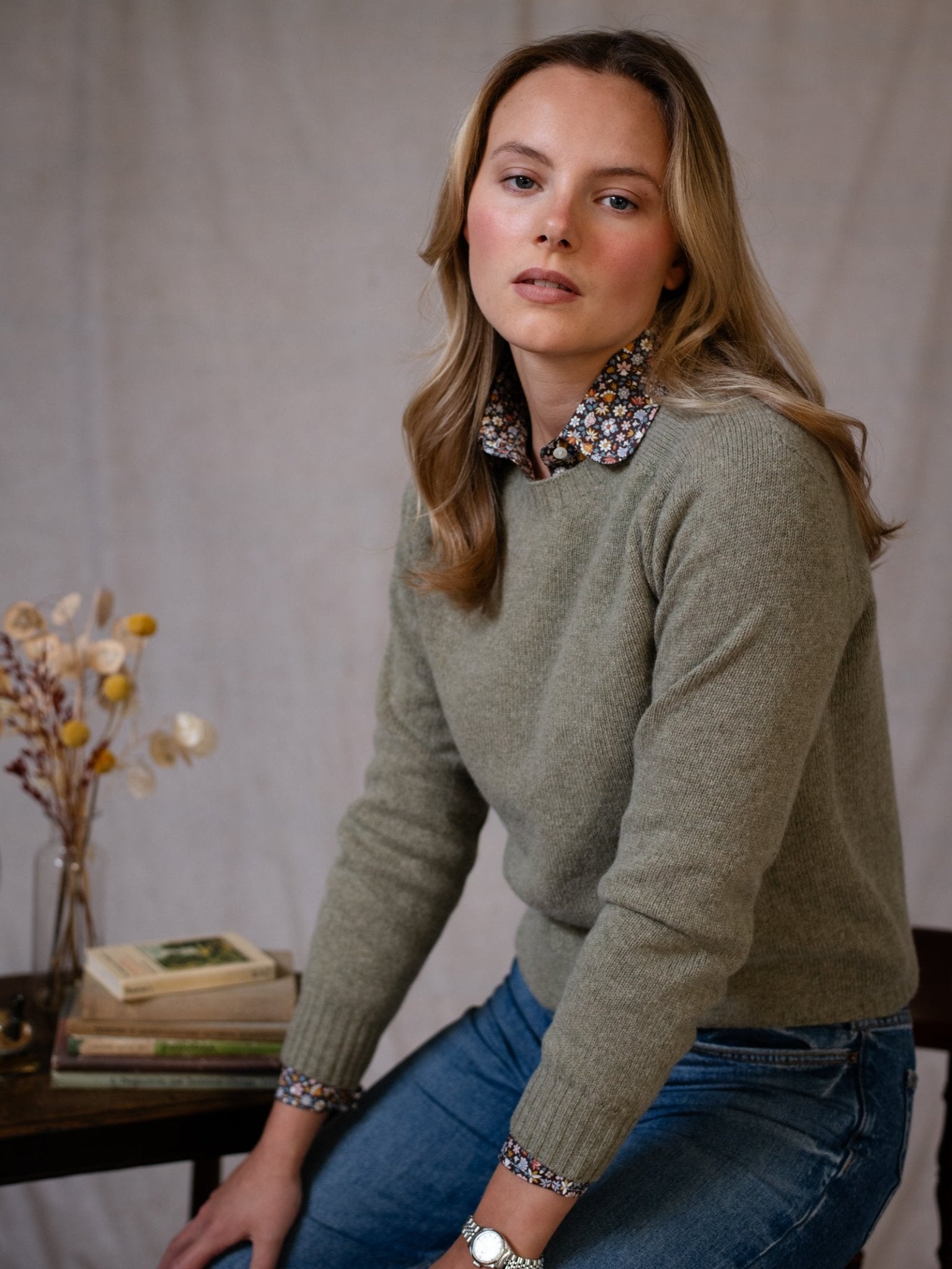 A woman with long blonde hair wears the Campbell's of Beauly Geelong Lambswool Crew Neck Jumper over a floral collared shirt and blue jeans, sitting on a table by books and dried flowers against a neutral backdrop.