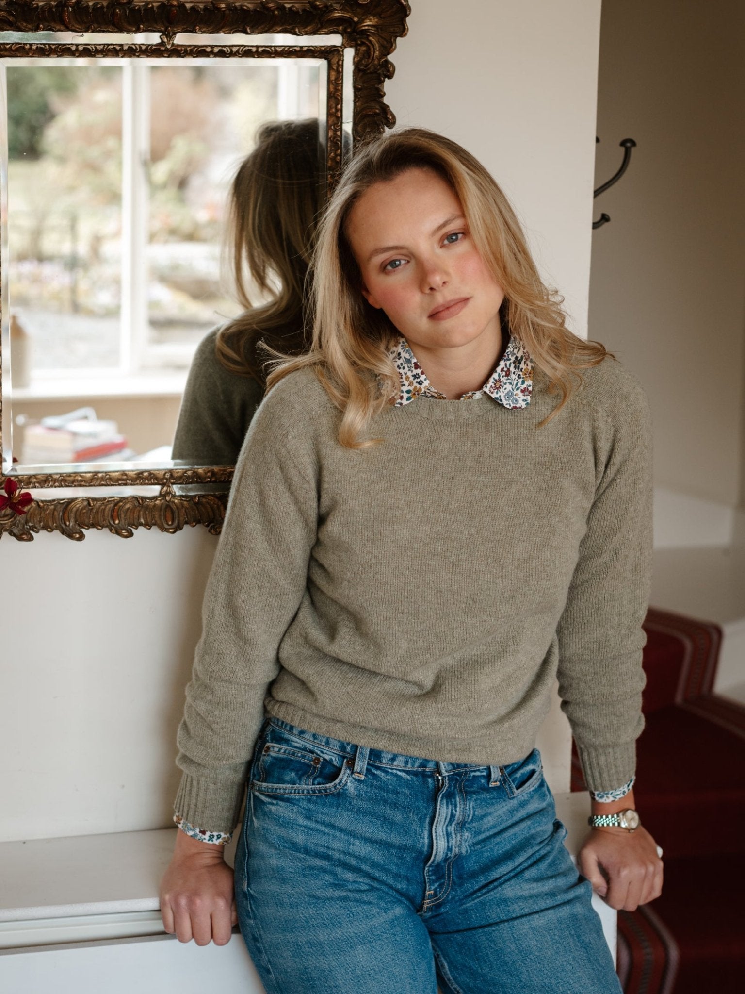A blonde woman in a beige Campbell's of Beauly Geelong Lambswool Crew Neck Jumper, floral-collared shirt, and blue jeans leans against a white wall in a cozy room with a framed mirror, calmly looking at the camera.
