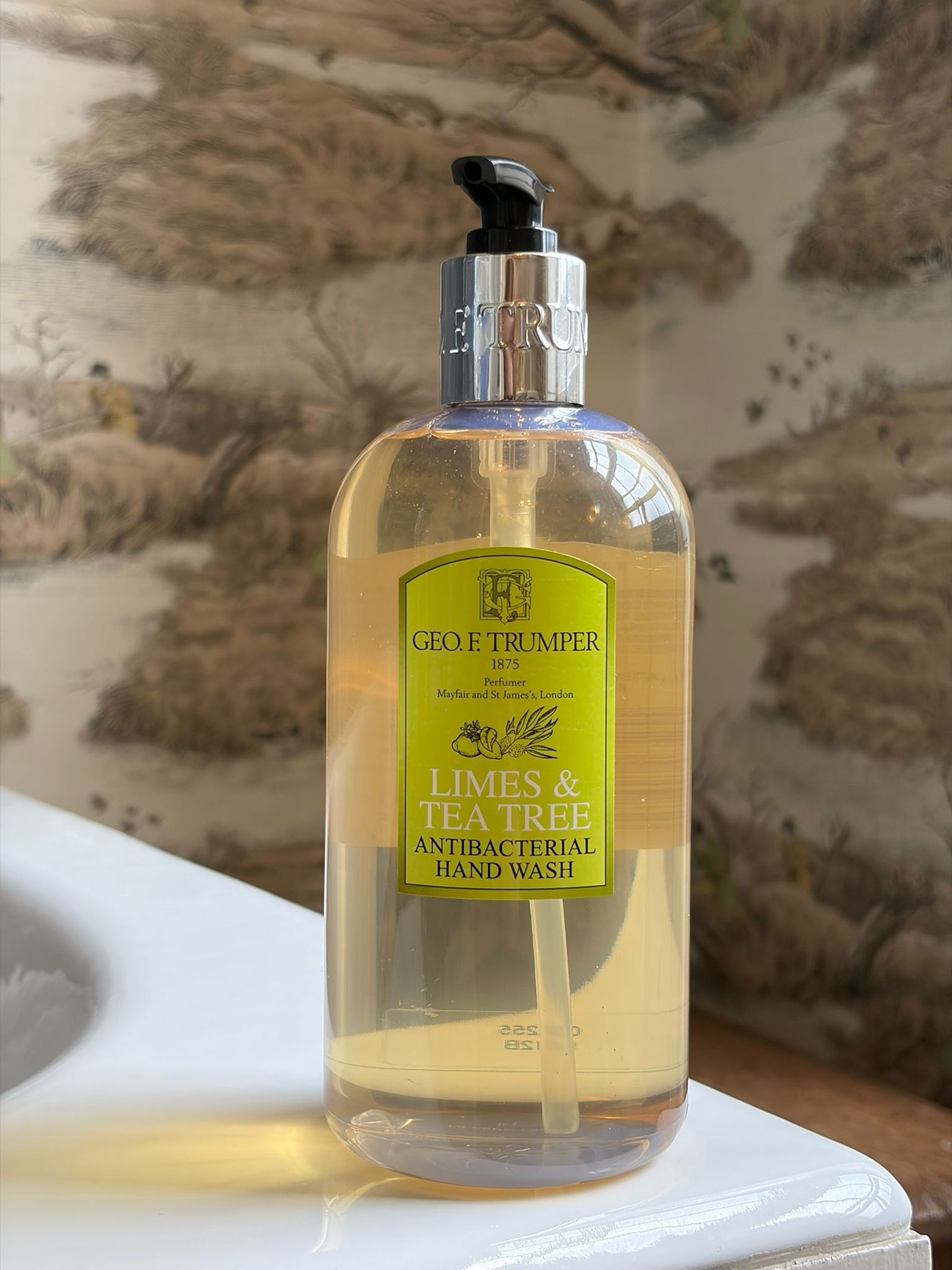 A clear pump bottle of Geo. F. Trumper Hair & Body Wash sits on a white sink, its vibrant label adding a touch of luxury against the nature-themed wallpaper background.