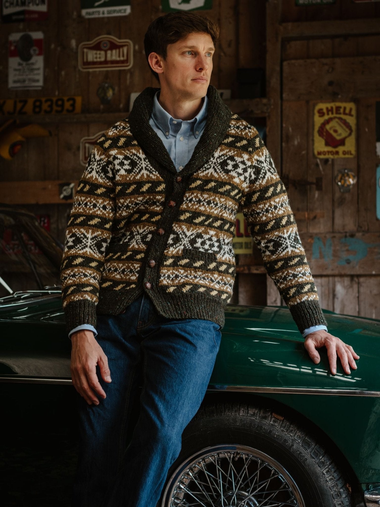 A man in a Hand Knit Fairisle Shawl Cardigan by Campbell’s of Beauly leans against a classic green car, with vintage signs and wooden walls in the background.