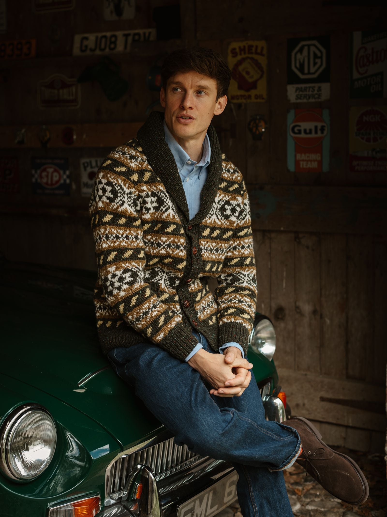 A man wearing the Campbell’s of Beauly Hand Knit Fairisle Shawl Cardigan in Olive Fairisle and jeans sits on the hood of a classic green car inside a rustic garage decorated with vintage signs.