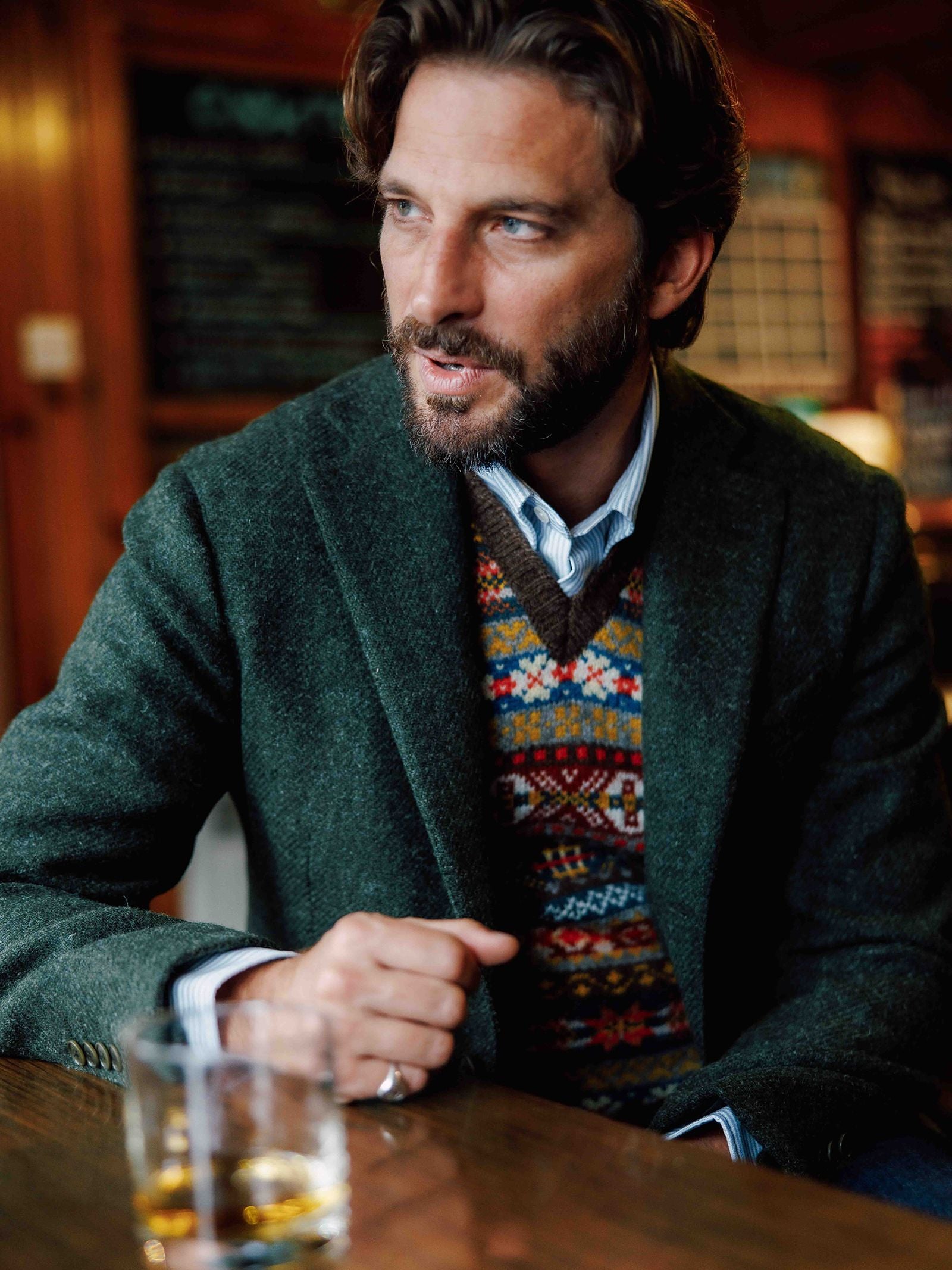 A bearded man sits at a wooden table in a cozy setting, wearing a regular fit Campbell’s of Beauly Harris Tweed Jacket and a colorful patterned sweater vest. A glass with a drink is in front of him, and chalkboards with writing can be seen in the background.