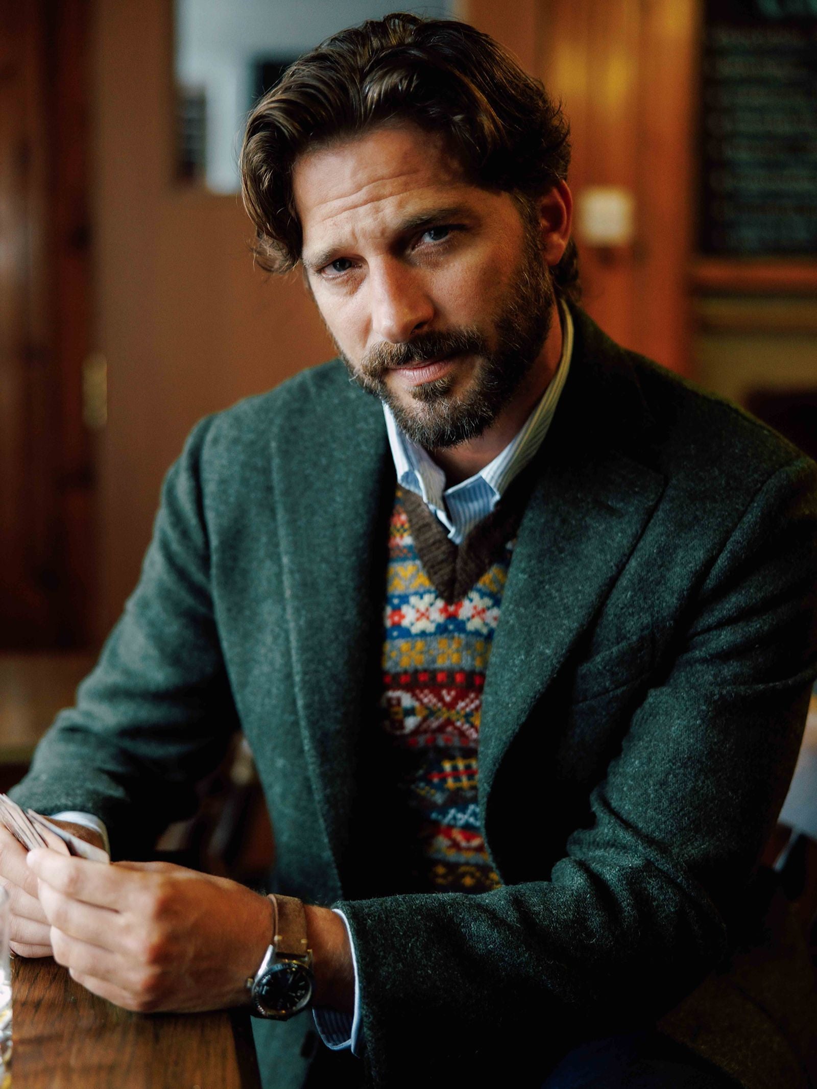 A man with wavy brown hair and a beard sits at a table wearing a stylish green Harris Tweed Jacket from Campbell’s of Beauly in a regular fit, paired with a patterned sweater vest and striped shirt, holding papers. The background is softly blurred in warm tones.
