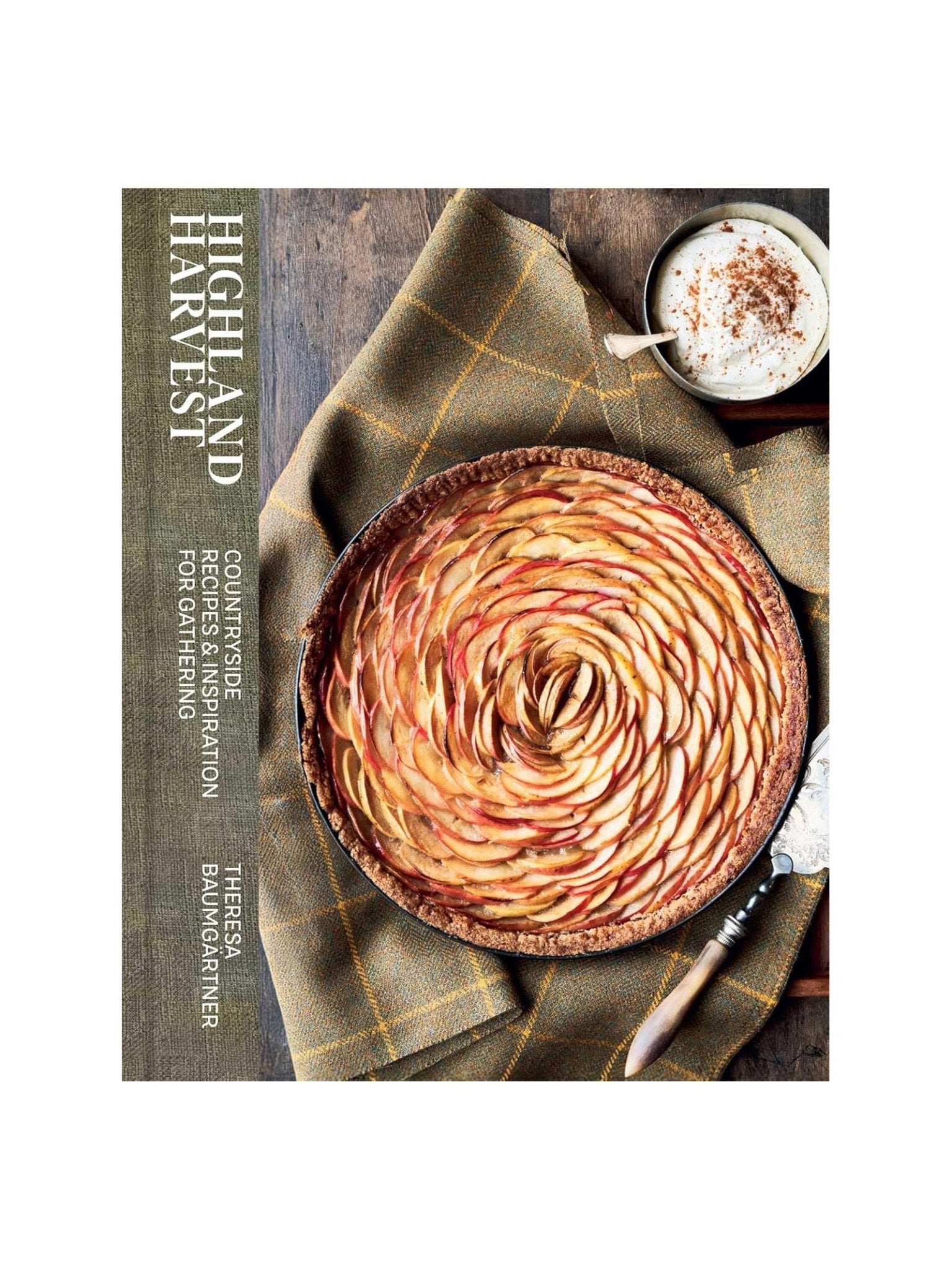 A rustic apple tart rests on a plaid cloth beside cinnamon whipped cream and a serving knife. Highland Harvest Book by Campbell's of Beauly, seen on the left, suggests delicious classic Scottish recipes inside.