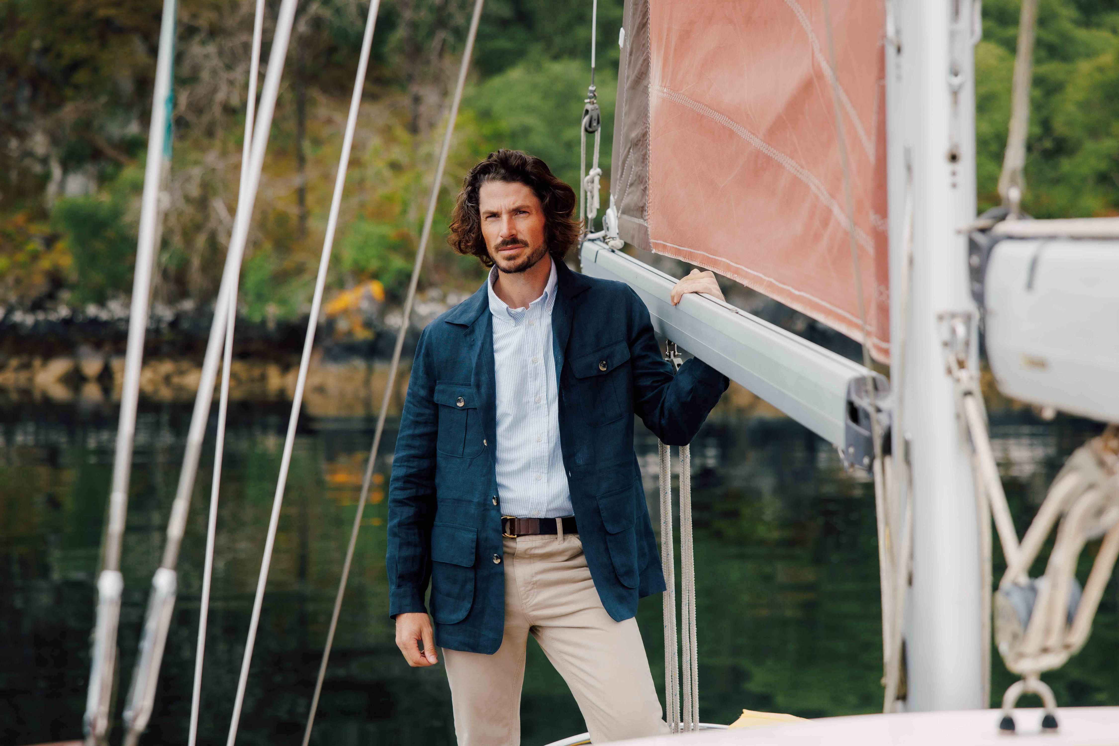 A man with shoulder-length brown hair, wearing a blue blazer and beige pants, stands on a sailboat holding the boom of a pink sail, with calm water and greenery in the background.