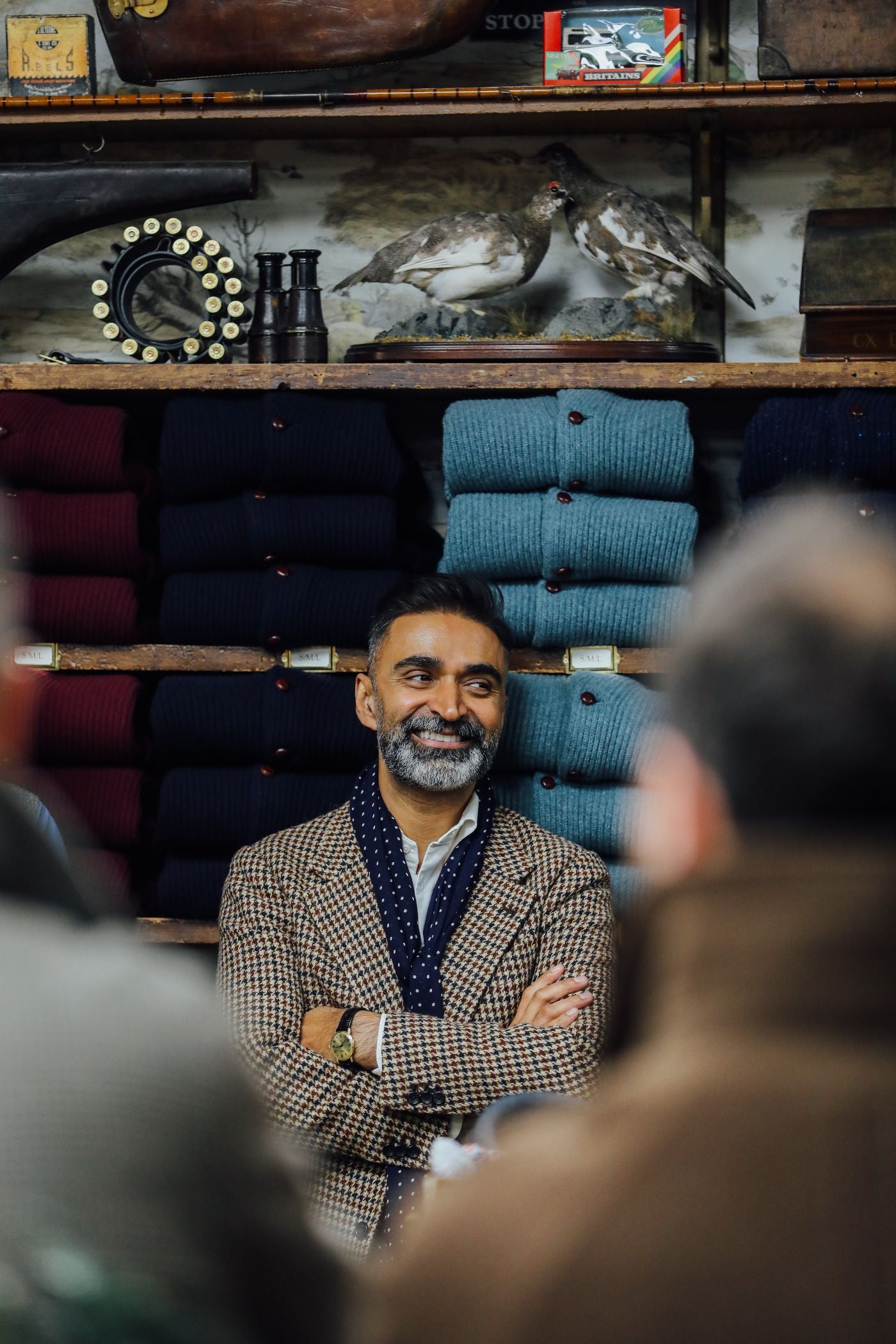 A man with a beard and mustache stands smiling with arms crossed in front of neatly stacked sweaters in various colors, with shelves and decorative items behind him. Two blurred figures are in the foreground.