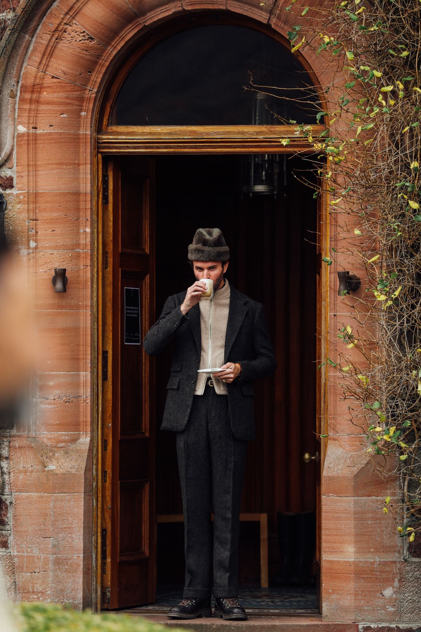 A man in a dark suit, hat, and cream scarf stands in a stone doorway, drinking from a cup and holding a plate. Climbing vines frame the entrance, and wooden doors are open behind him.