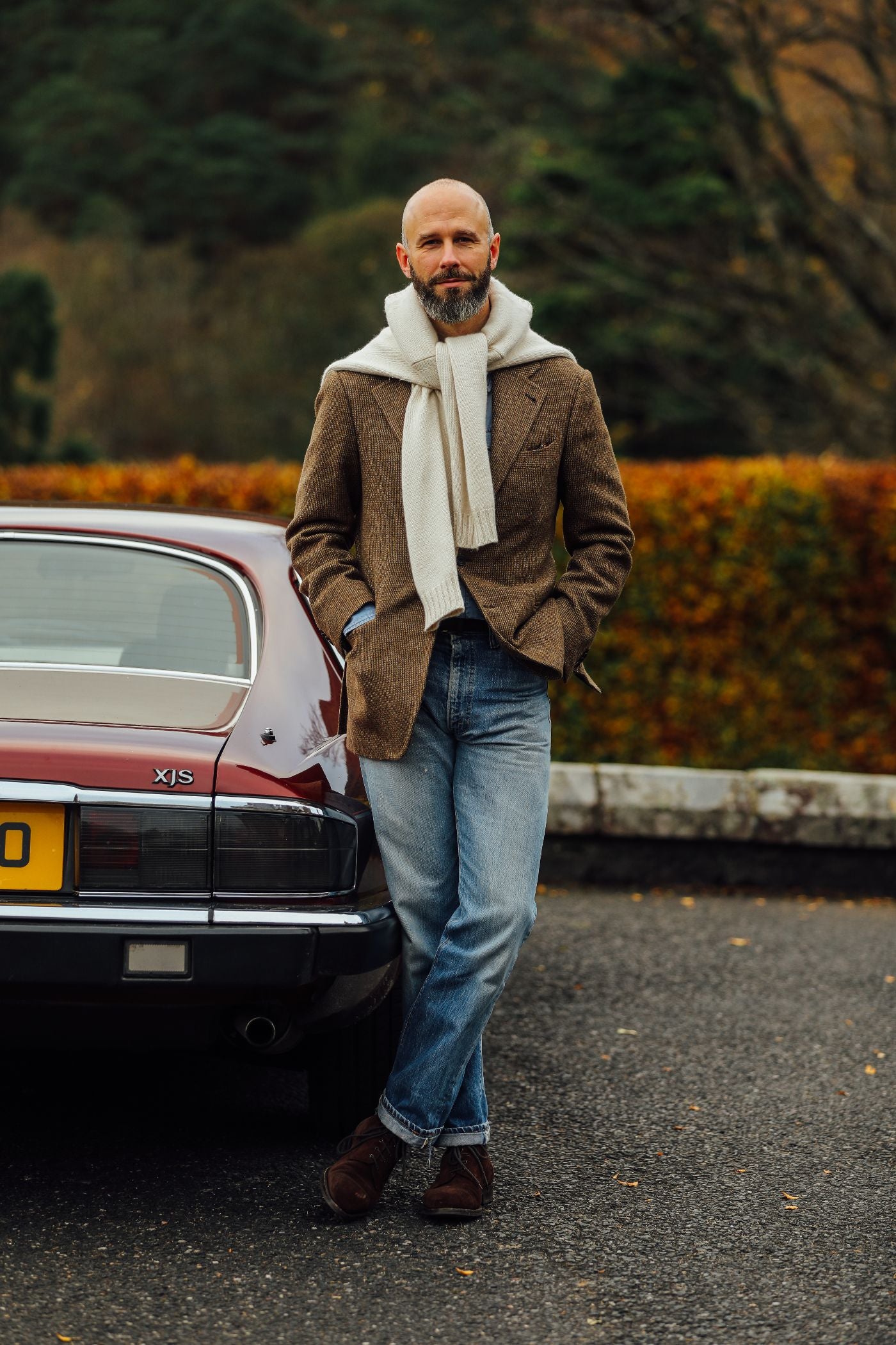 A man with a beard, wearing a brown blazer, blue jeans, brown shoes, and a white scarf, stands with hands in pockets, leaning against a maroon classic car parked by a hedge with autumn foliage.