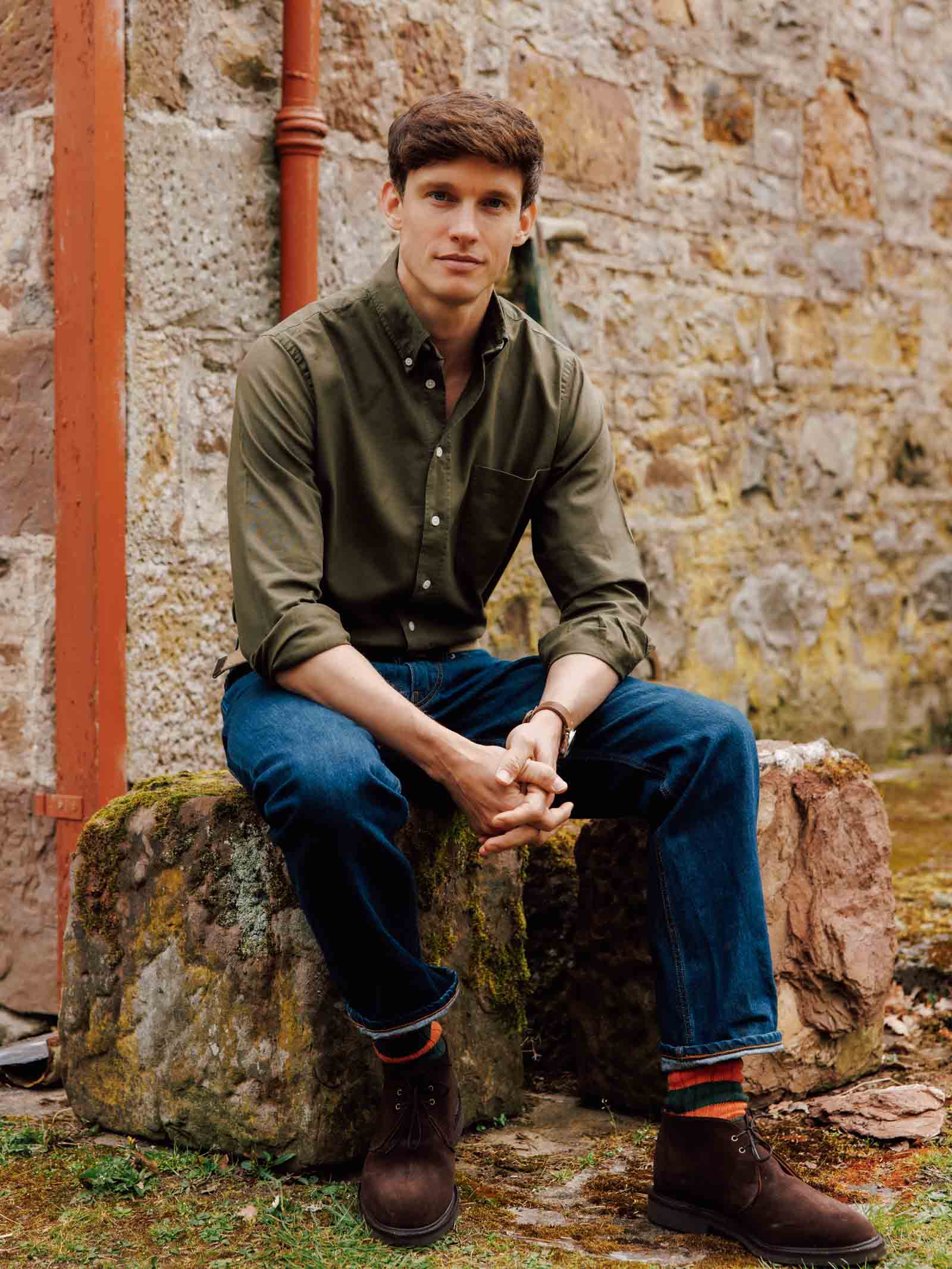 A young man wears a Campbell's of Beauly Oxford Button Down Shirt in green and blue jeans as he sits on a mossy stone bench outdoors, hands clasped and legs crossed, in front of a weathered stone wall.