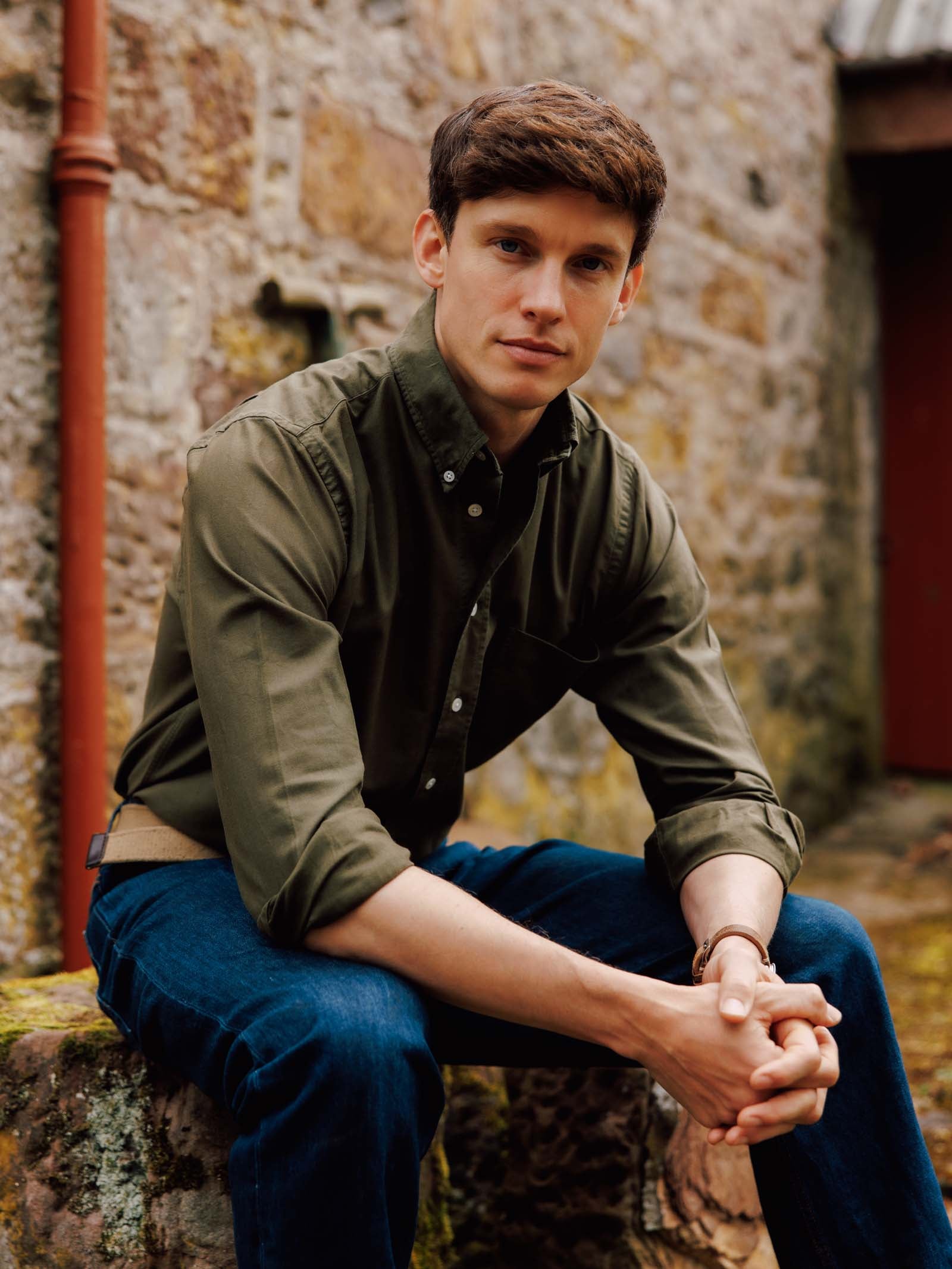 A young man with short brown hair, wearing the Campbell's of Beauly Oxford Button Down Shirt and blue jeans, sits on a stone ledge in front of a rustic wall, looking calmly at the camera with his hands clasped.