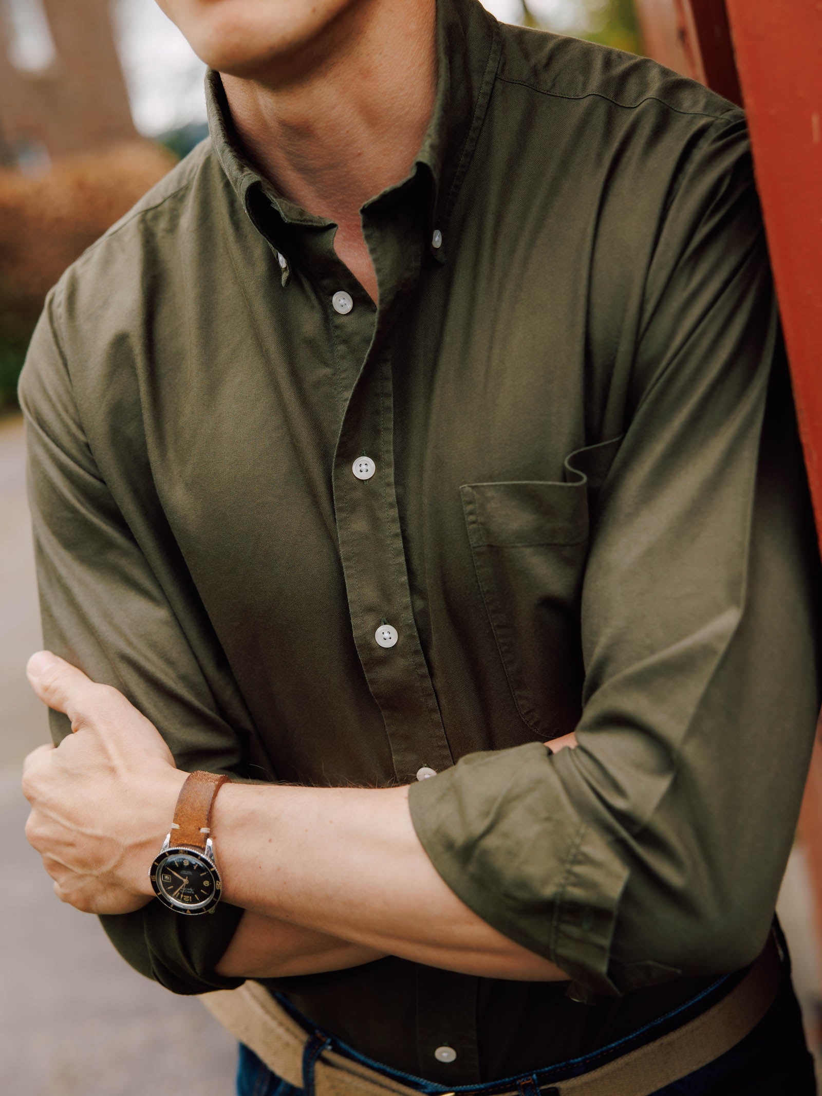 A person in a green Campbell's of Beauly Oxford Button Down Shirt and a brown leather wristwatch stands with arms crossed, leaning on a red wall. Their face is mostly out of frame.