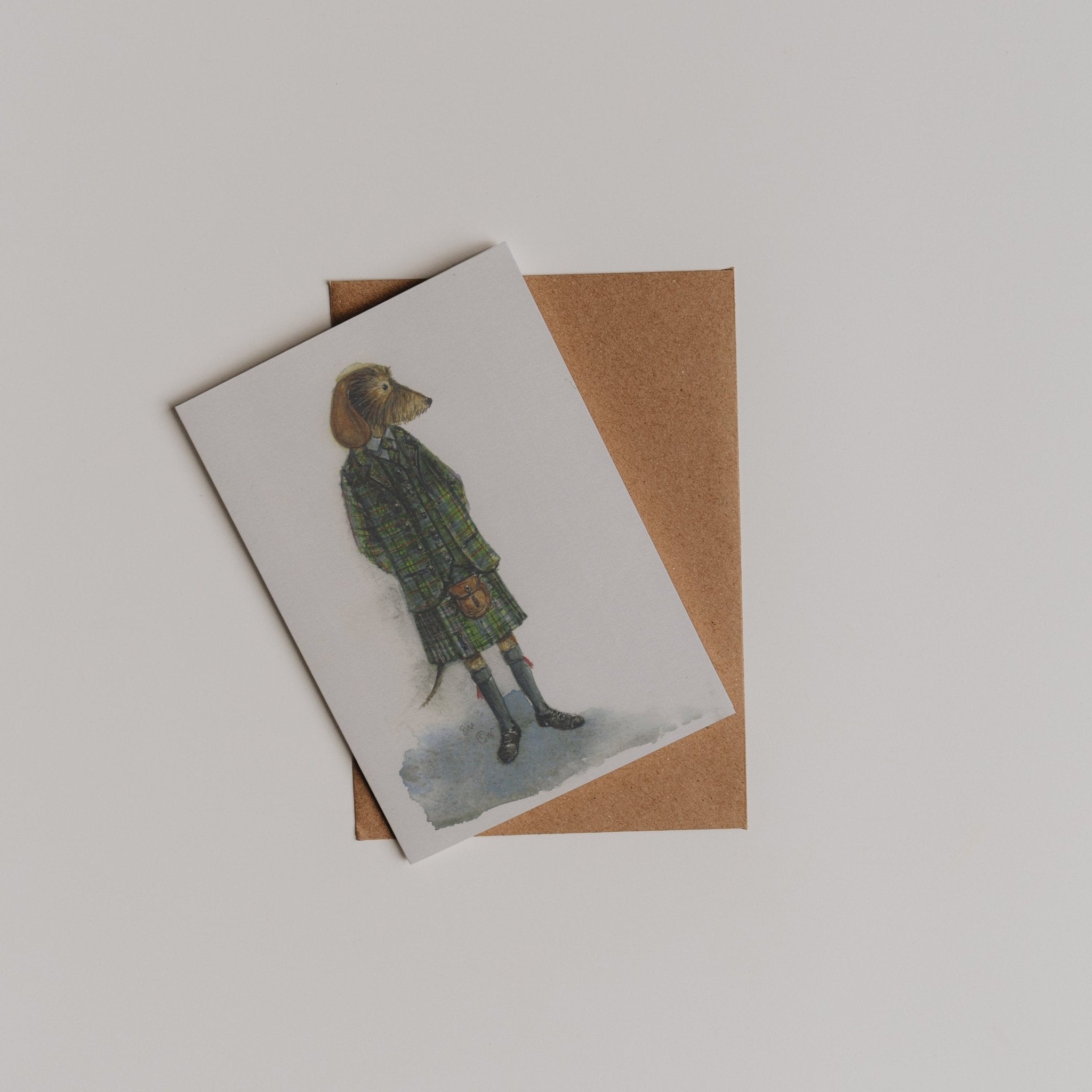 The Kilted Dachshund Greeting Card by Campbell's of Beauly features an illustrated dog in a green plaid coat, shown atop a brown envelope on a plain white background.