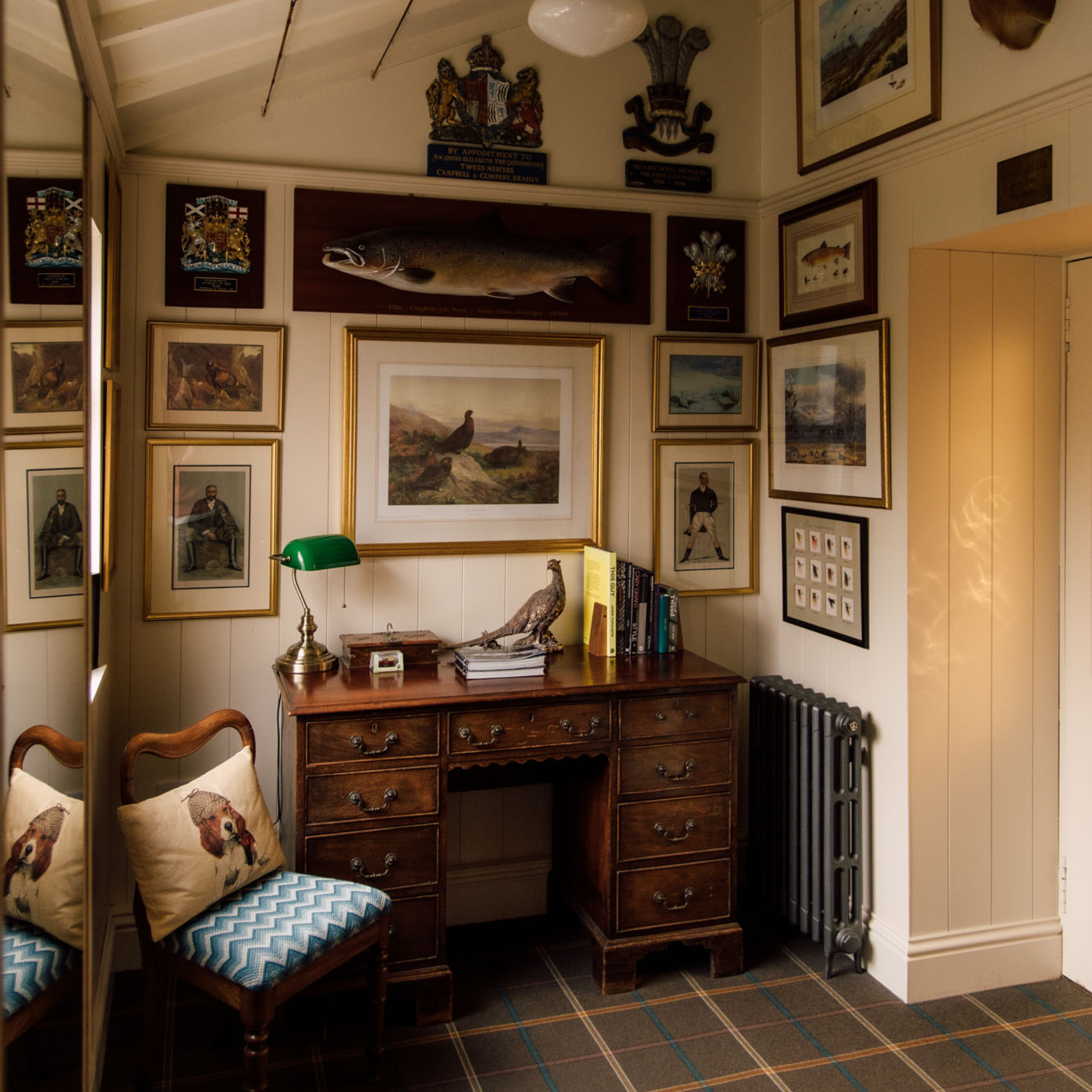 A cozy study corner with a wooden desk, books, a green desk lamp, and a bird sculpture. Surrounding walls display framed artwork, crests, and a mounted fish above the desk. Two cushioned chairs sit nearby.