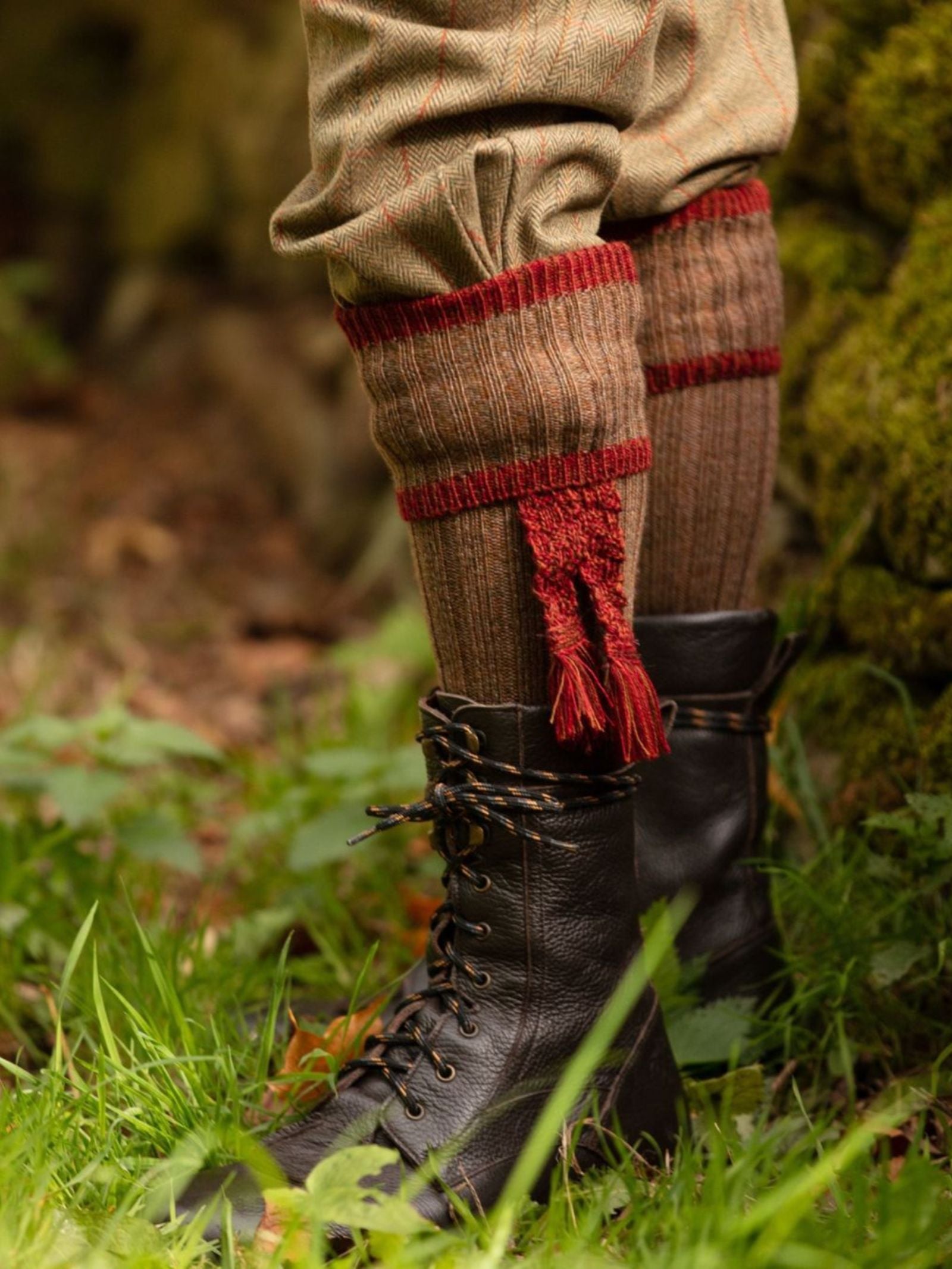 A close-up of a person’s lower legs wearing Campbell's of Beauly Kyle Socks—brown with red cuffs and tassels—paired with tweed trousers and black lace-up boots, standing on green grass outdoors.