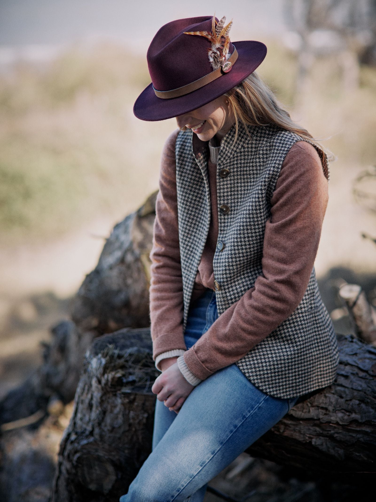 Wearing a Hicks & Brown Auld Stock Gamebird Fedora with feather wrap, checked vest, pink sweater, and blue jeans, a person sits on a fallen tree outdoors, looking down with hands clasped.