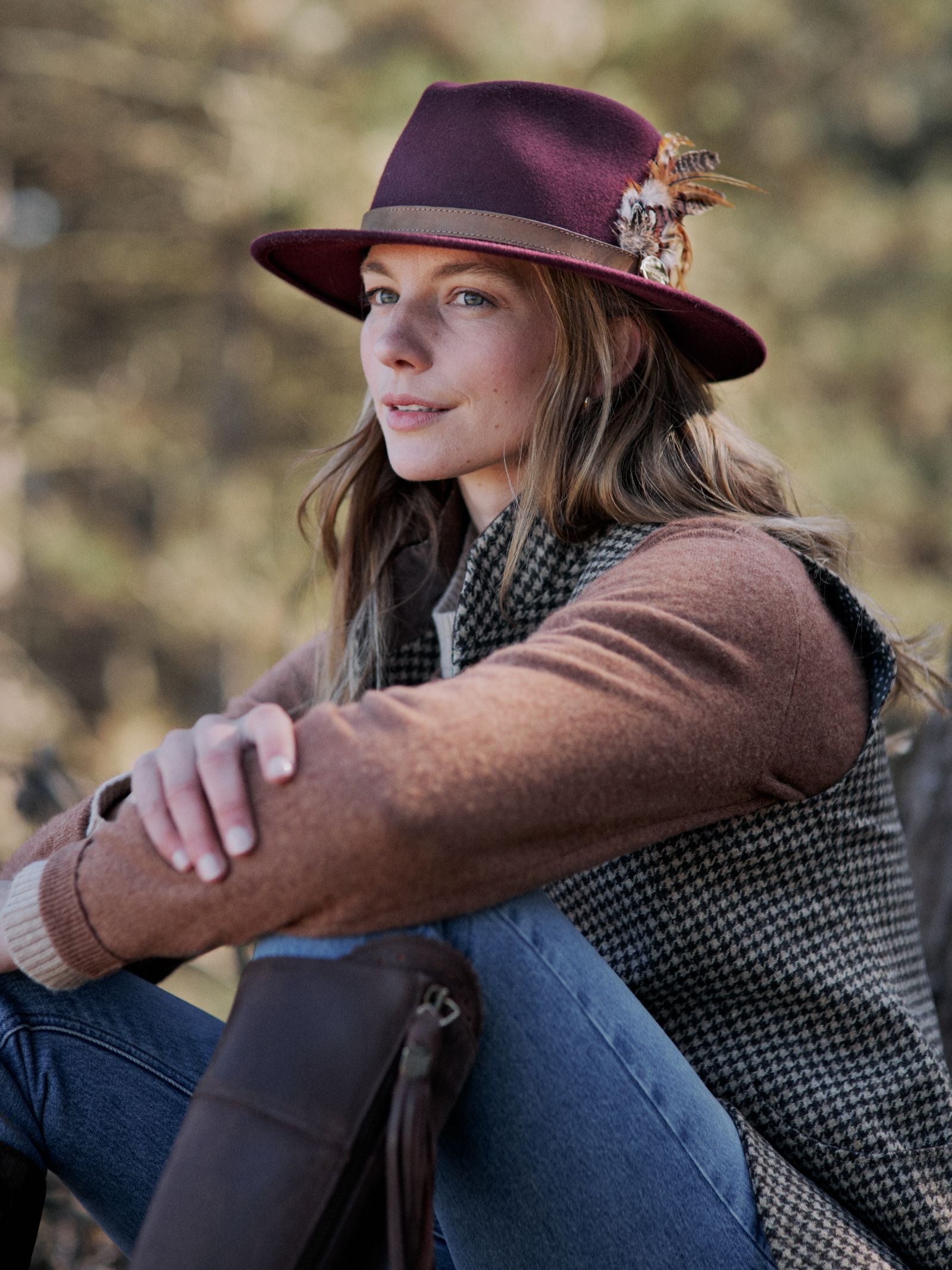 A woman sits outdoors, wearing the Hicks & Brown Auld Stock Gamebird Fedora with decorative feathers, a brown sweater, and a houndstooth vest. She gazes calmly to the side against a softly blurred background of greenery.