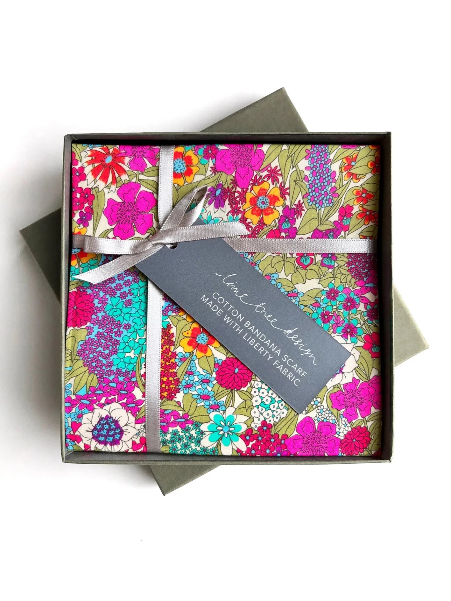 The Campbell's of Beauly Square Headscarf, made from vibrant floral Liberty cotton, comes folded in a stylish gift box with a white ribbon and a tag reading "Cotton Bandana Scarf Made with Liberty Fabric." It’s a versatile way to add color to any look.