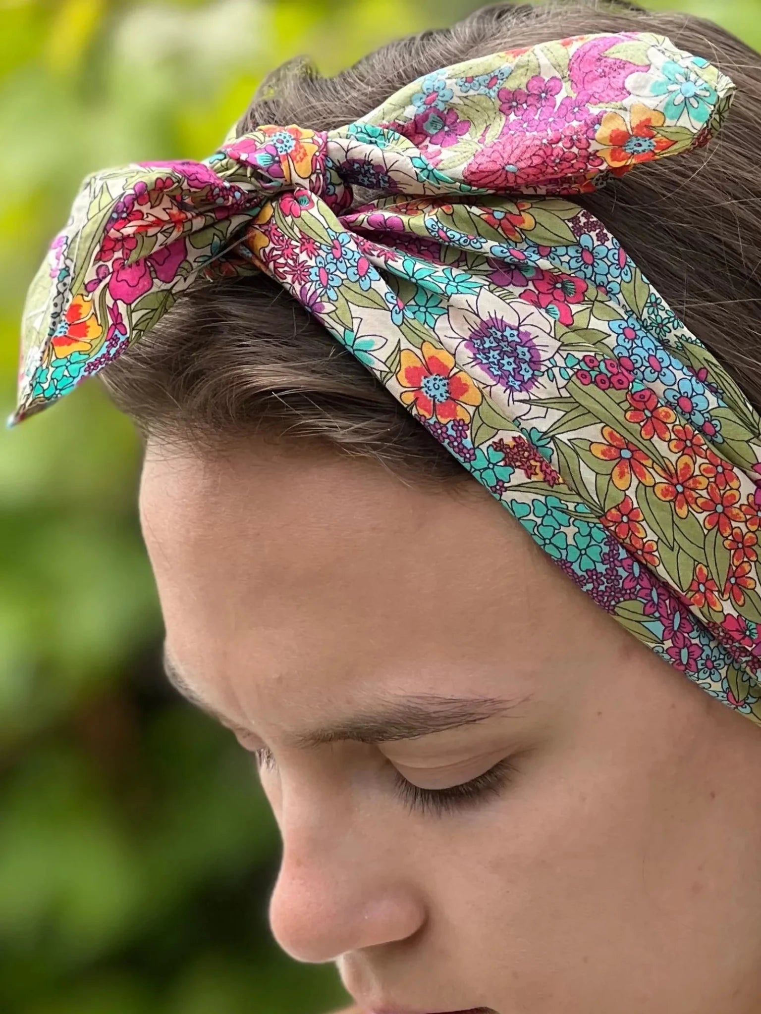 A close-up of a person in profile wearing the Campbell's of Beauly Square Headscarf with a colorful floral pattern, tied as a headband in a bow. Blurred greenery forms the background.