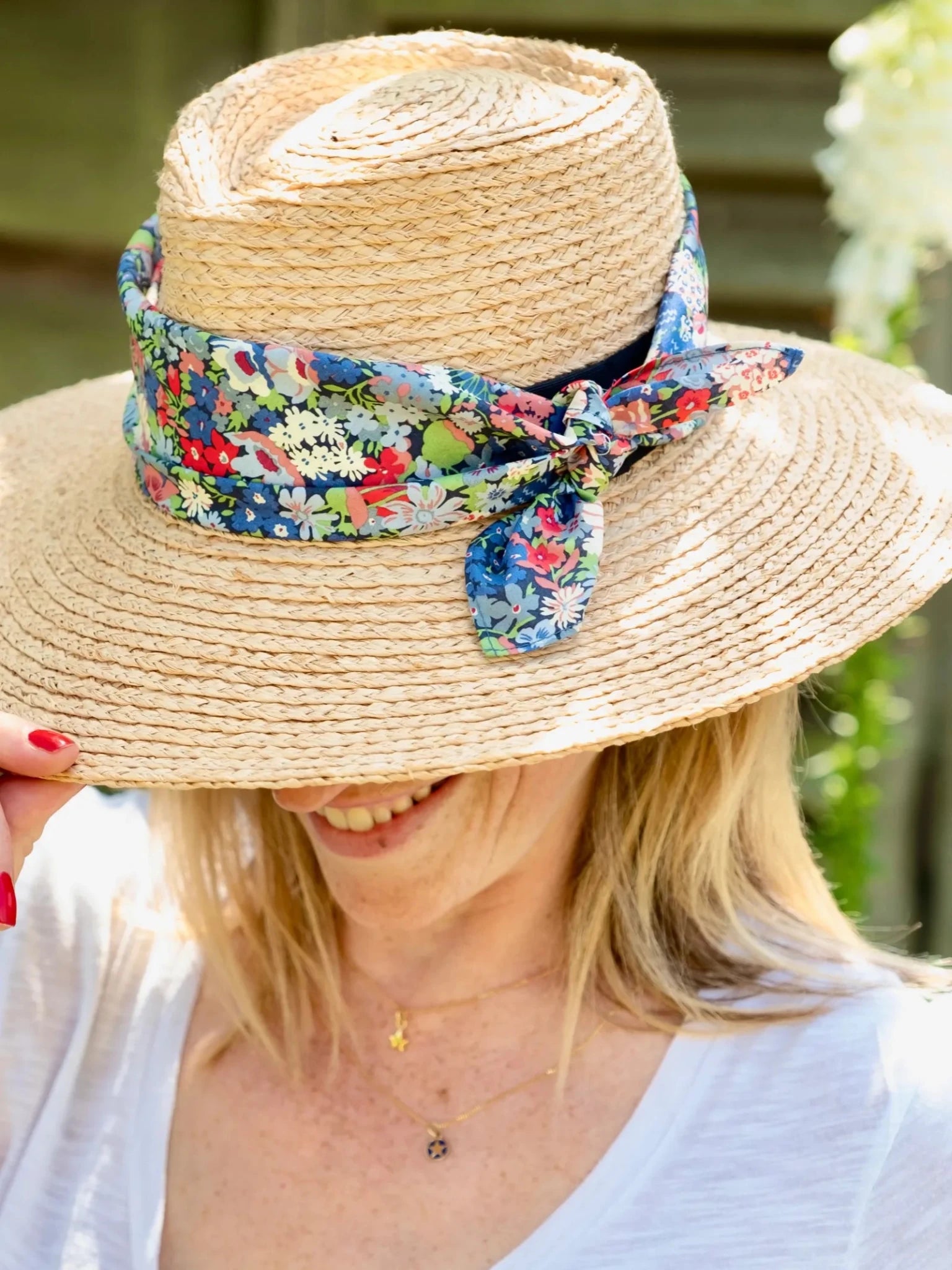 A blonde woman smiles in a wide-brimmed straw hat adorned with the Square Headscarf by Campbell's of Beauly. Her white top and the partially obscured face add to her elegant, stylish look.