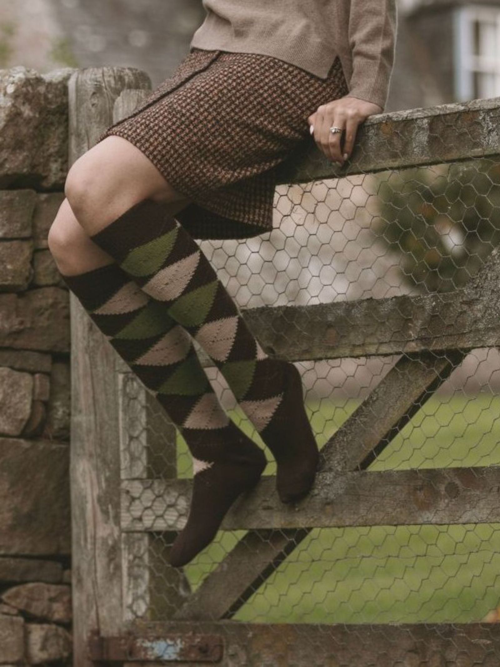 Wearing Campbell's of Beauly Argyle Socks, a person sits outdoors on a wooden gate by a stone wall, dressed in a brown sweater and patterned skirt.