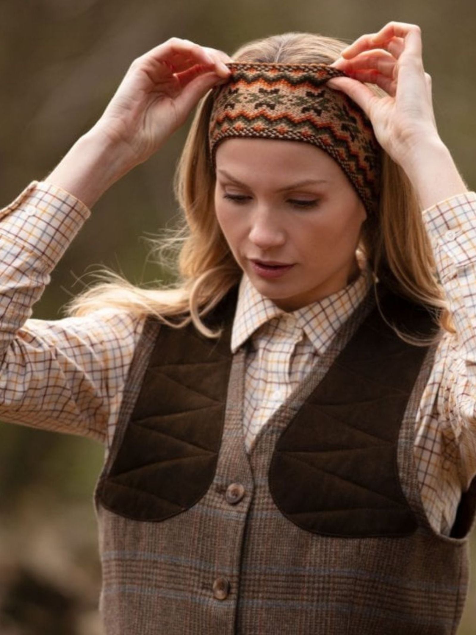 A woman outdoors in the scenic Scottish Borders adjusts a Campbell's of Beauly Fairisle Headband, crafted from 100% wool, over her forehead. She wears a plaid shirt and brown vest, with a blurred natural background behind her.