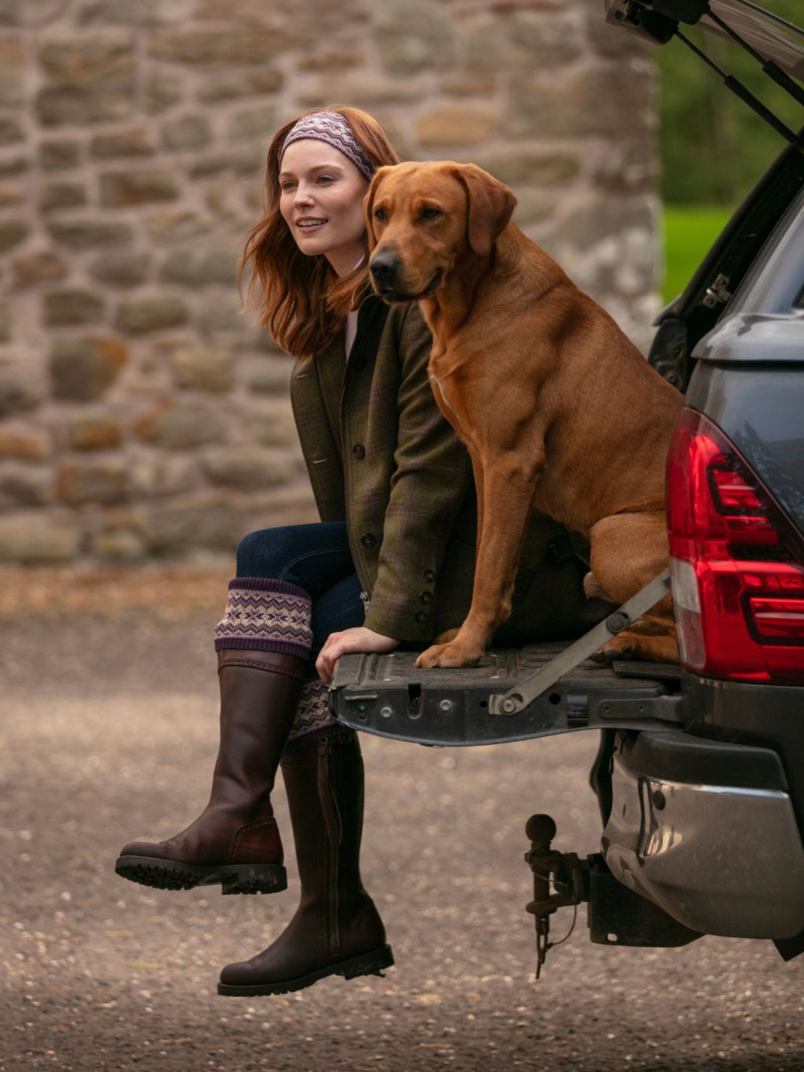A woman wearing the Campbell's of Beauly Fairisle Headband sits on a vehicle’s tailgate with her large brown dog, both smiling outdoors in the Scottish Borders in front of an old stone wall.