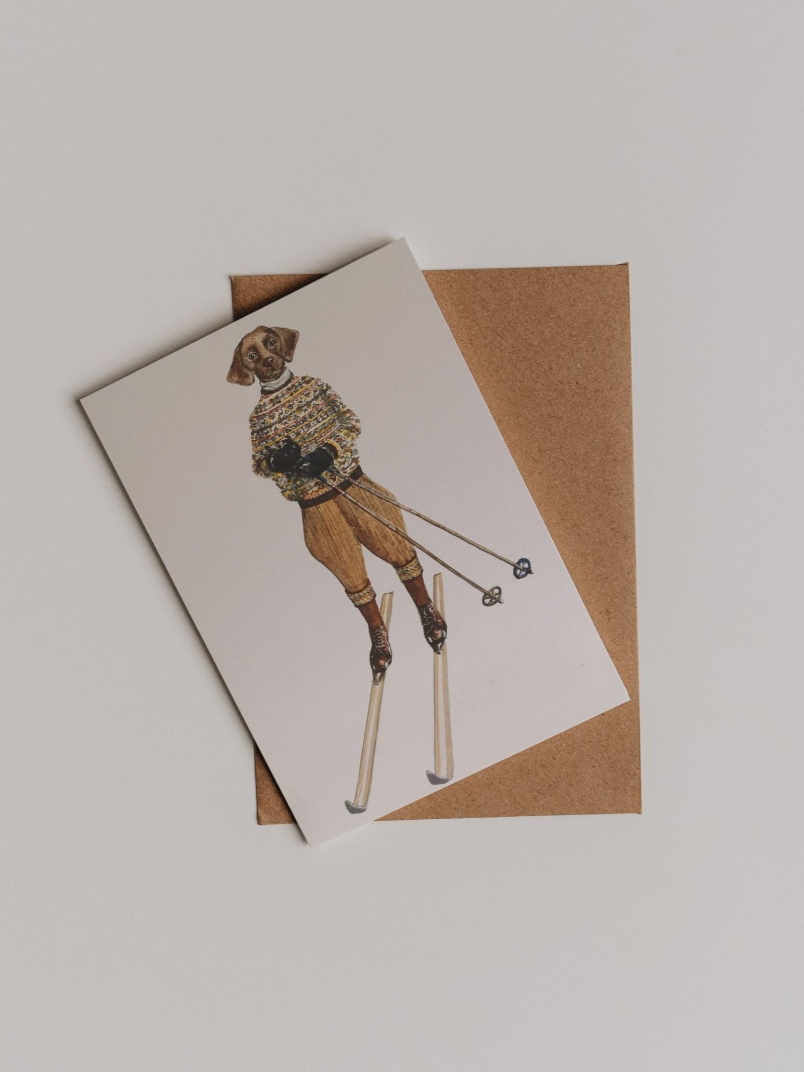 The Le Ski Greeting Card by Campbell’s of Beauly features a Sophie Murray-inspired dog in vintage ski attire and skis, set atop a brown envelope against a white background.