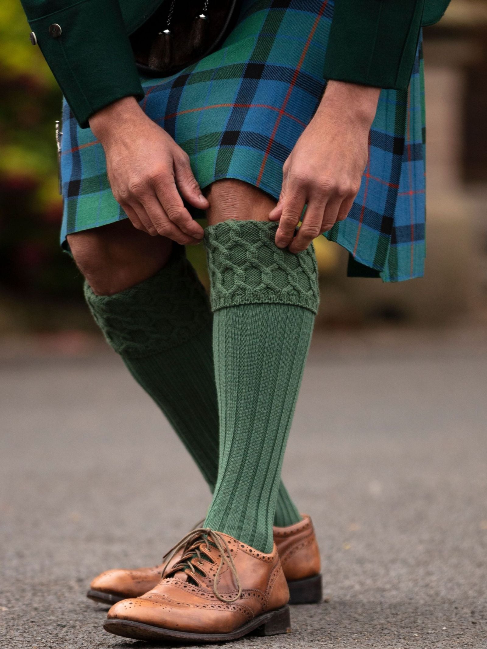 A person in Highland dress, wearing a blue and green tartan kilt, adjusts their Campbell's of Beauly Kilt Socks and brown leather brogues while standing on a paved surface.