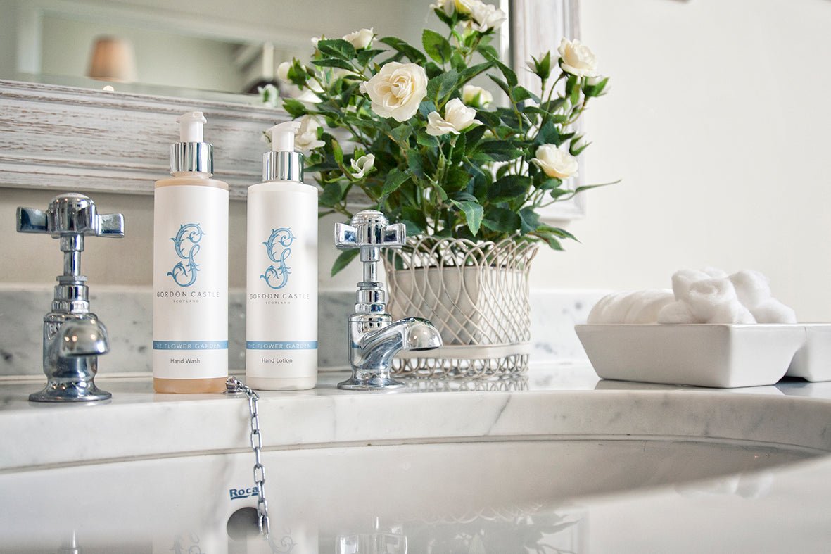 A bathroom sink with silver faucets, two bottles of hand wash, Gordon Castle’s The Flower Garden Hand Cream, a potted white rose plant, and a dish of cotton balls on a marble countertop in front of a mirror.