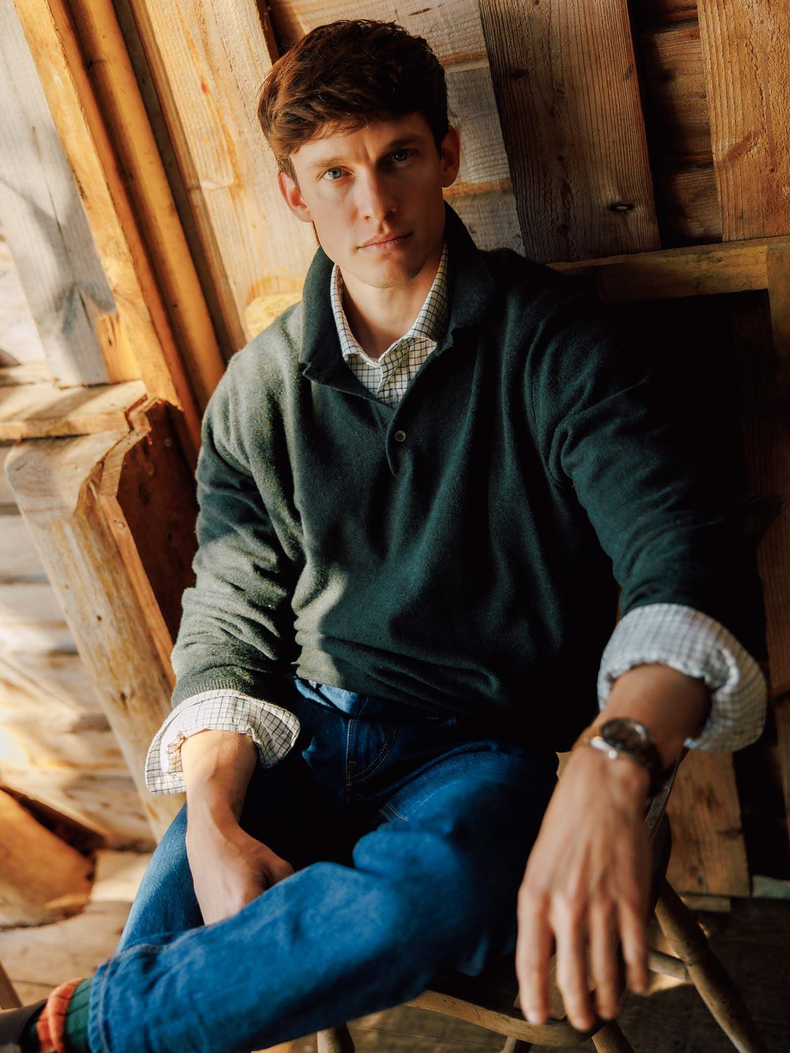A young man in a dark Campbell’s of Beauly Merino & Cashmere Rugby sweater and jeans sits on a rustic wooden chair, looking at the camera. Sunlight illuminates the tailored silhouette of his Italian knitwear and casts a warm glow on the wooden wall behind him.
