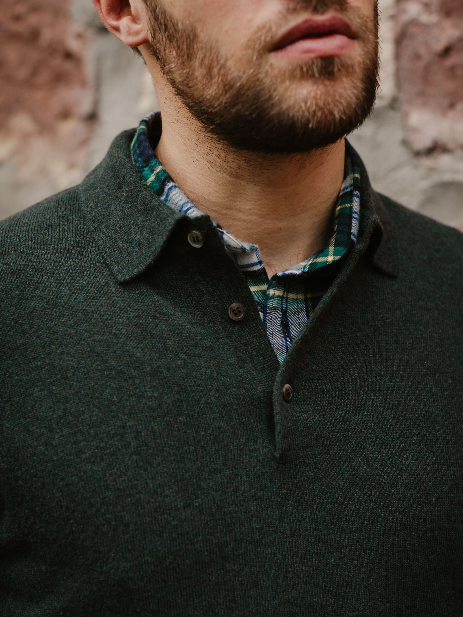 A man with a short beard wears the Campbell's of Beauly Merino & Cashmere Rugby sweater over a green and blue plaid shirt, standing before a textured brick wall. Only his lower face and upper torso are visible.